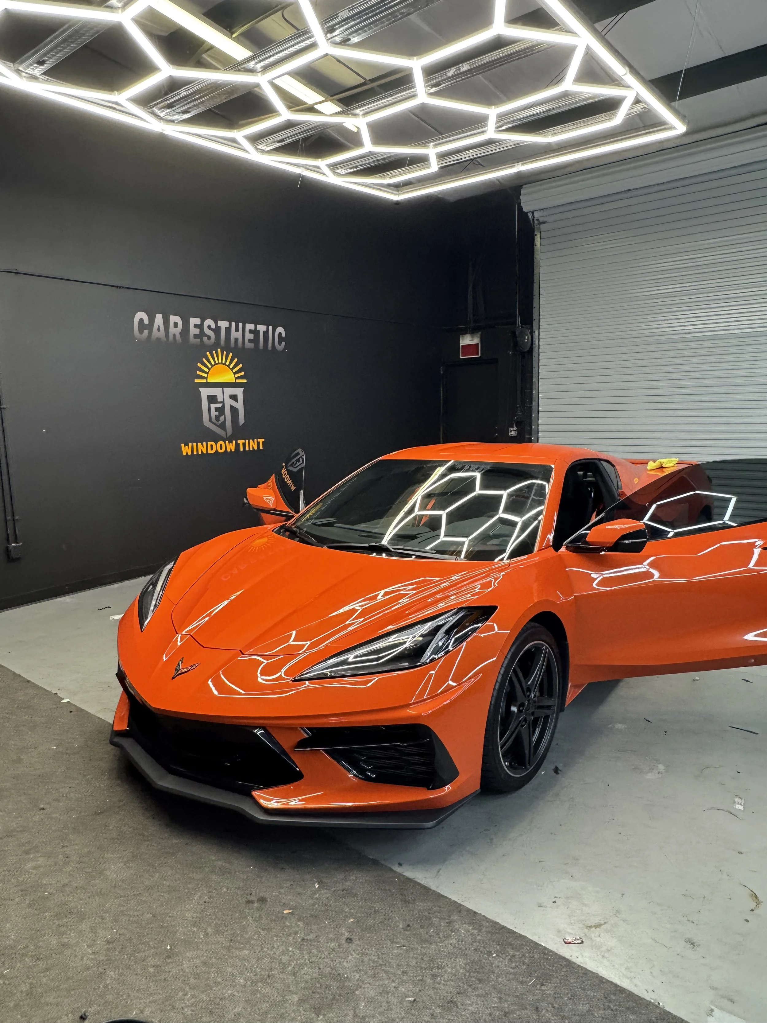 An orange sports car, a Chevrolet Corvette, parked inside a showroom with a black wall and a gray roll-up door. The ceiling features geometric light fixtures.