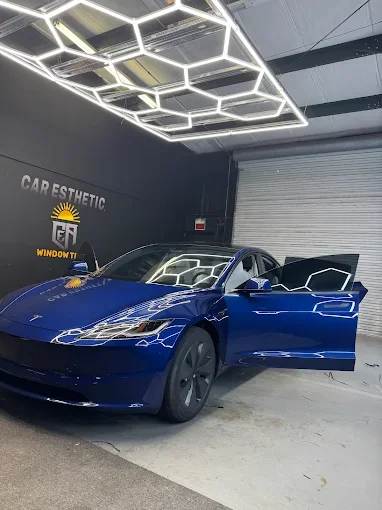 A blue Tesla sports car parked inside a modern garage with hexagonal LED light fixtures on the ceiling and a black wall with yellow branding.