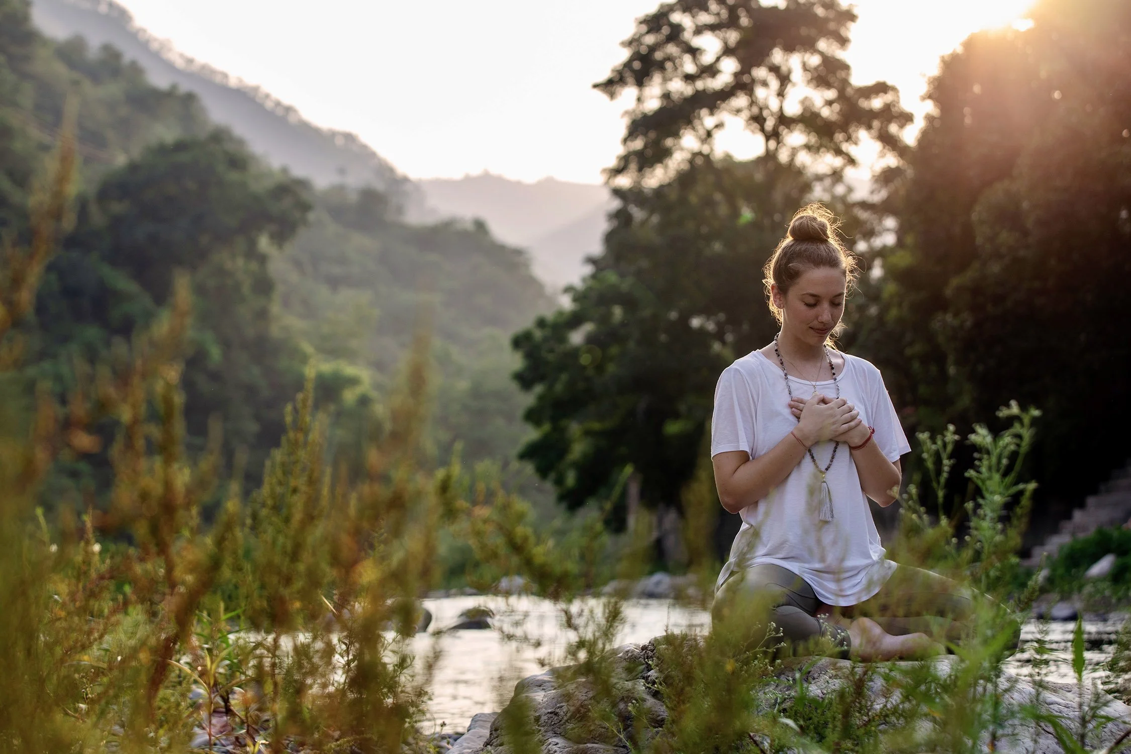 Girl seated in meditation posture with hands to heart in white shirt with mala beads next to Ganga river tributary in India