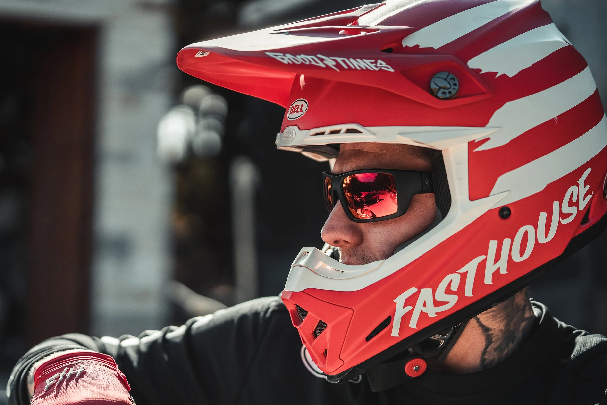 Close-up of a person wearing a red and white full-face helmet with brand logos and the word 'FASTHOUSE' visible, along with black sunglasses, as they look to the side.