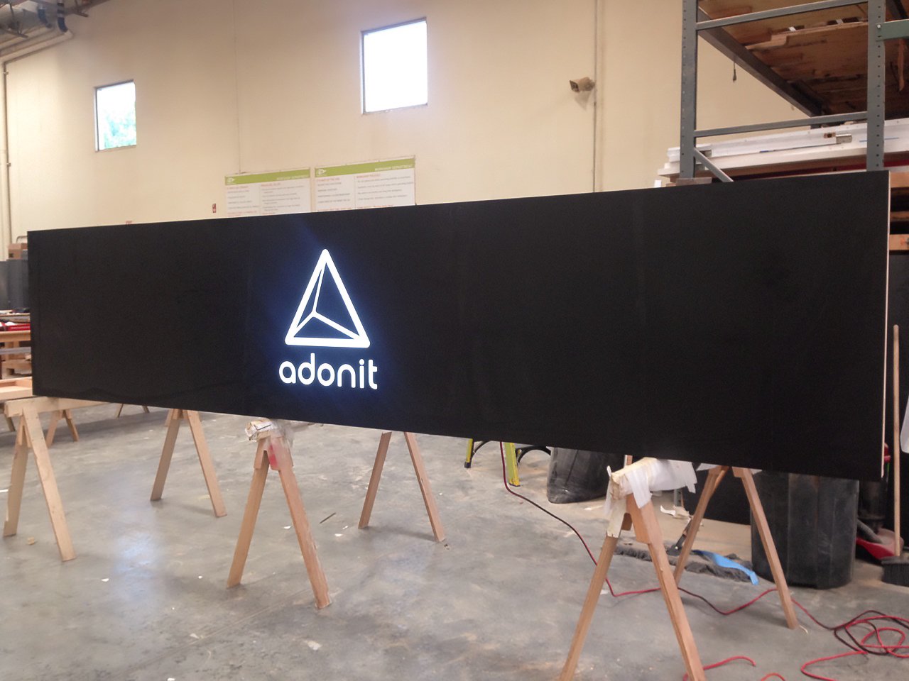 A large black illuminated sign with a white geometric logo and the word 'adonit' beneath it, displayed in a workshop or storage area on wooden supports.