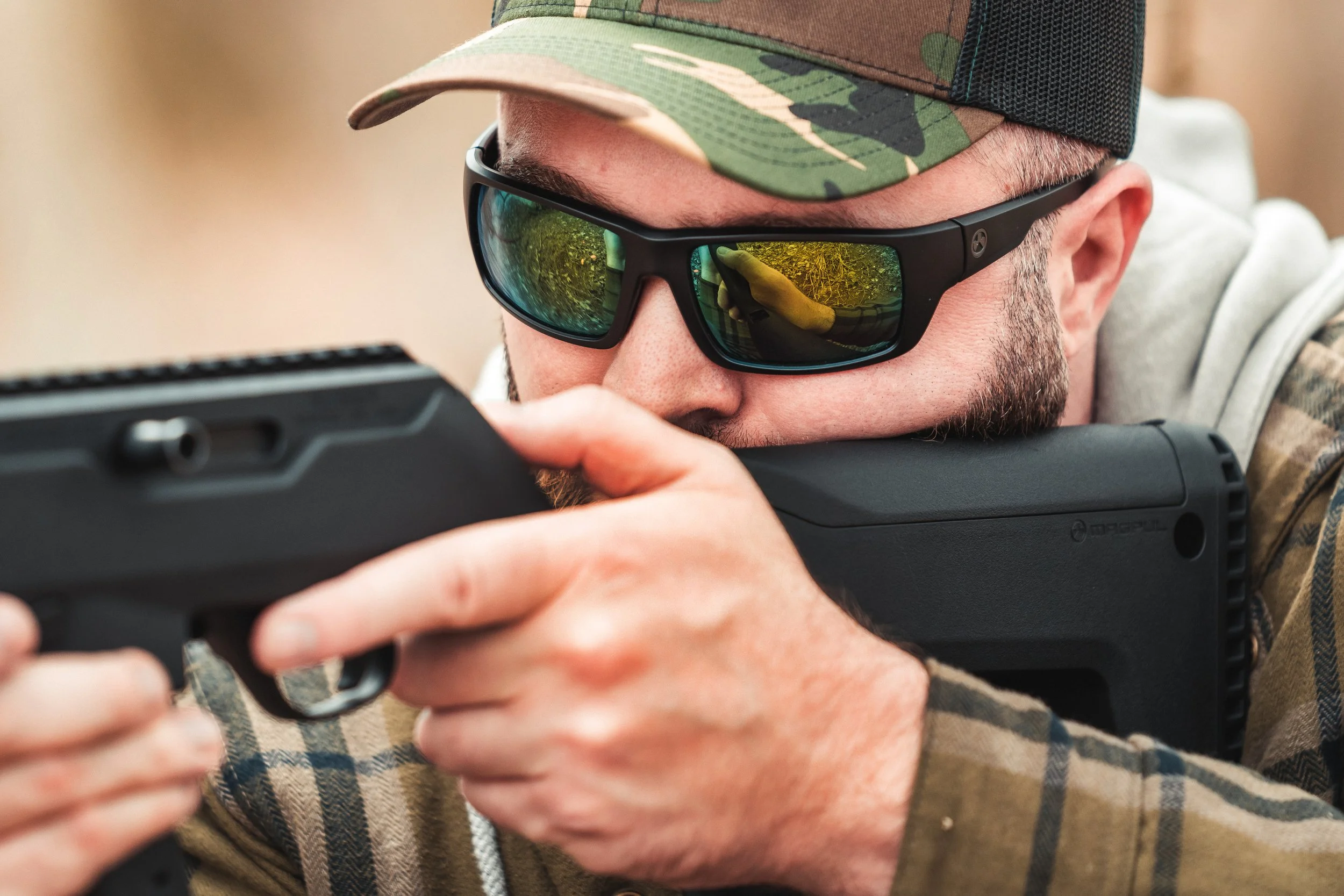 A person in camouflage cap and sunglasses aiming a black firearm, with reflection in the sunglasses.