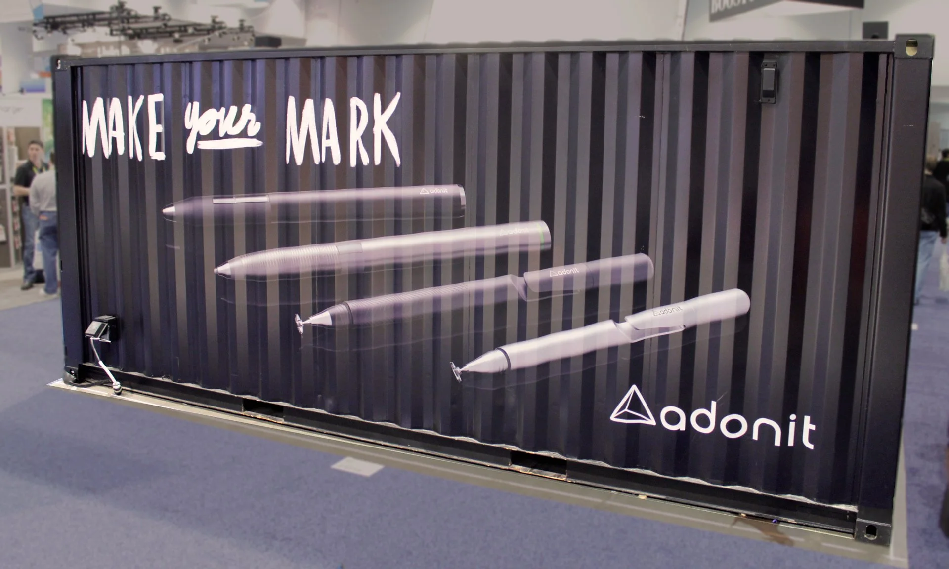 A display at CES conference with a mural showing four digital Adonit styluses with the phrase 'Make your mark' written above in white text. The display features the Adonit logo at the bottom right corner.