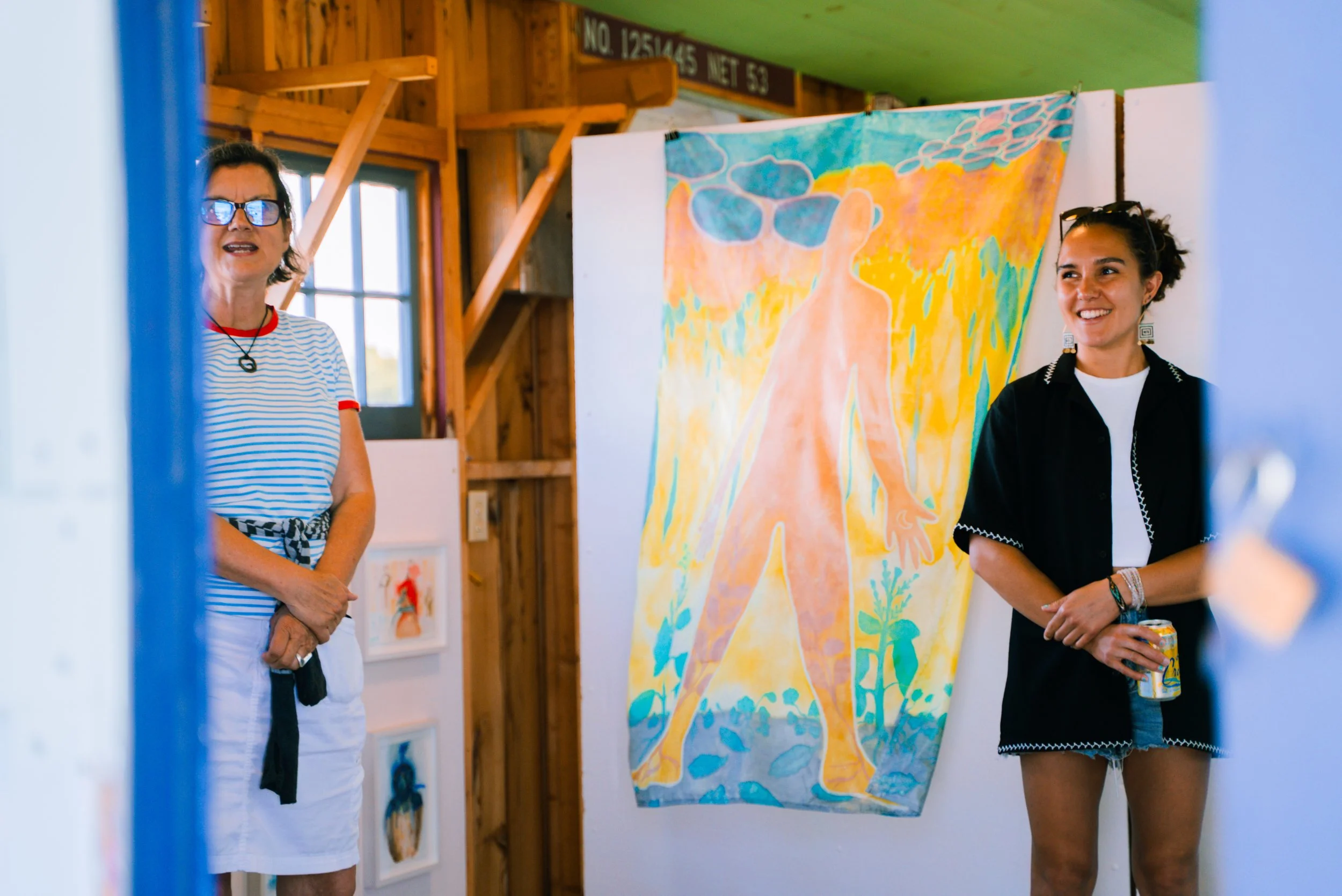 Two women standing in an art exhibit with a colorful silhouette painting on the wall. One woman is wearing sunglasses, a striped shirt, and a white skirt, while the other is holding a can and wearing a black shirt and denim shorts.