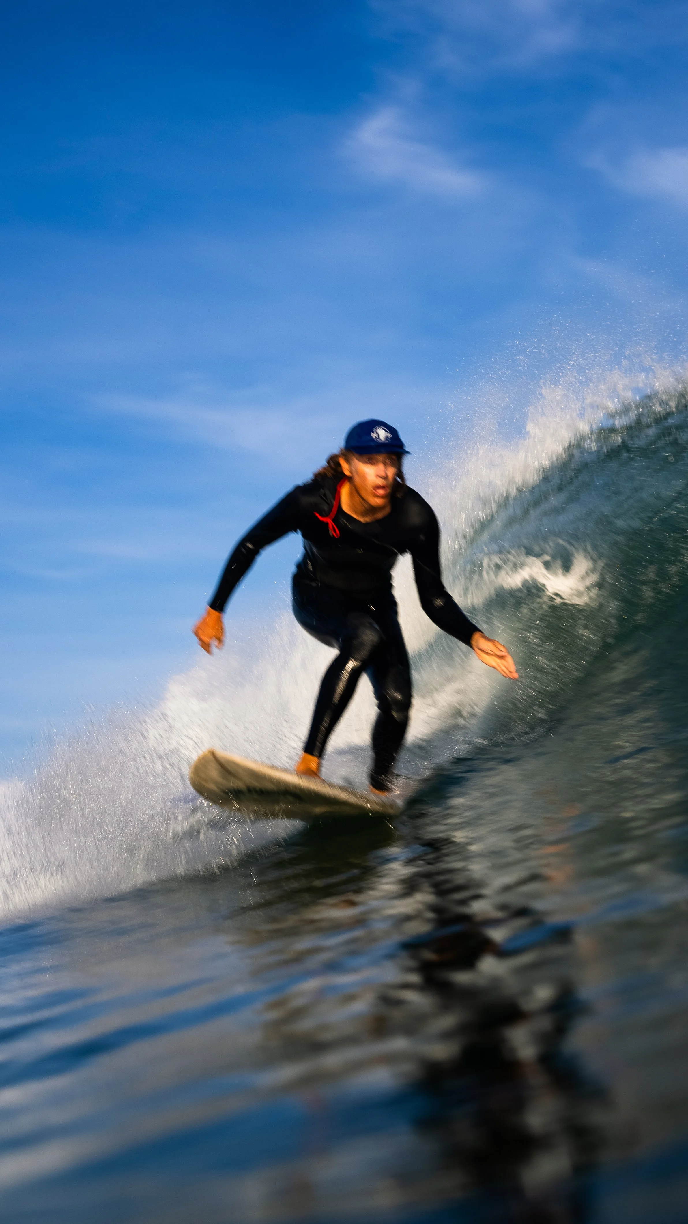 Surfer riding a wave wearing a wetsuit and cap, with ocean and blue sky in the background.