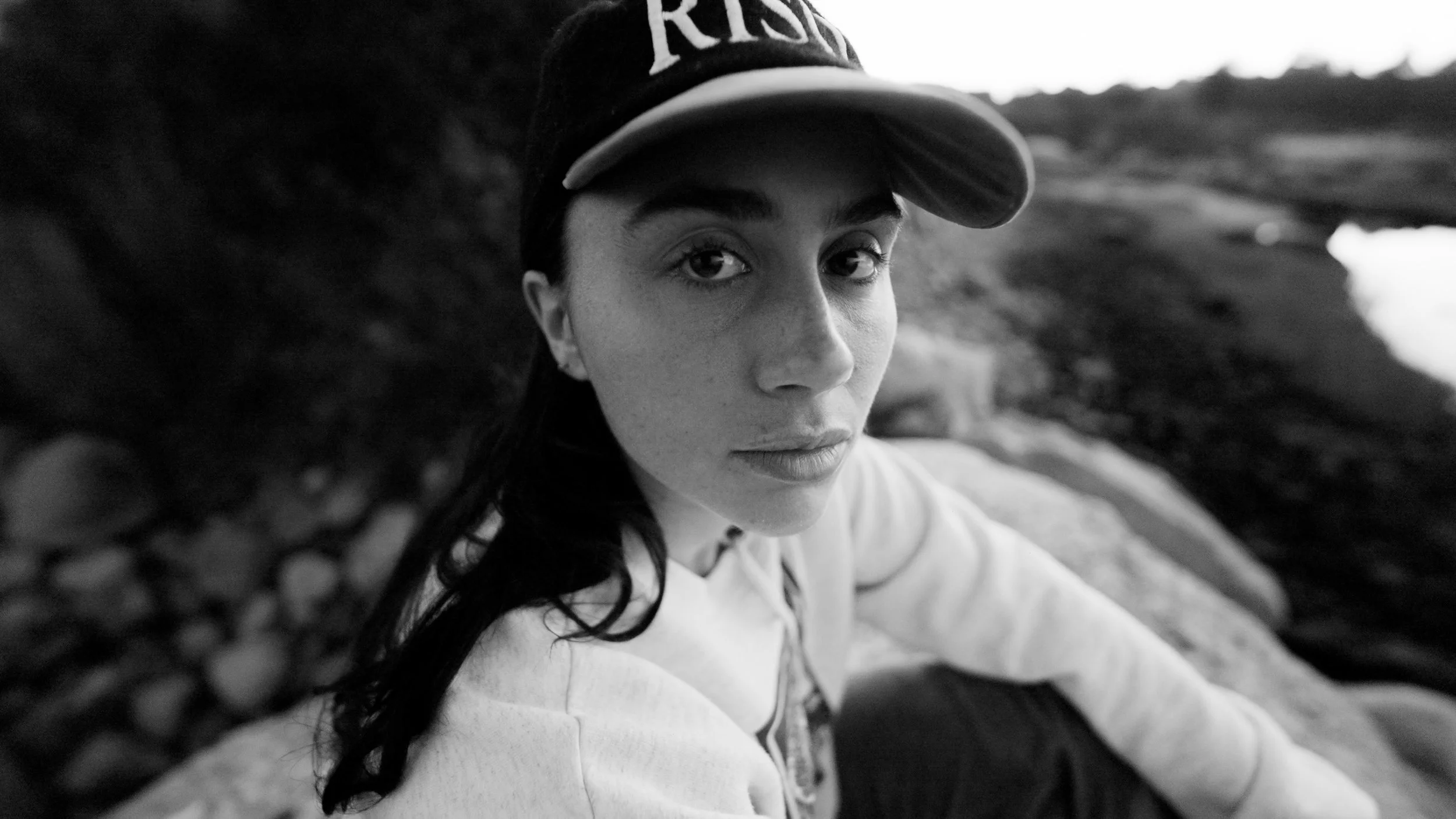 Black and white photo of a person wearing a cap with the word 'RISE,' sitting outdoors, with a blurred natural background.