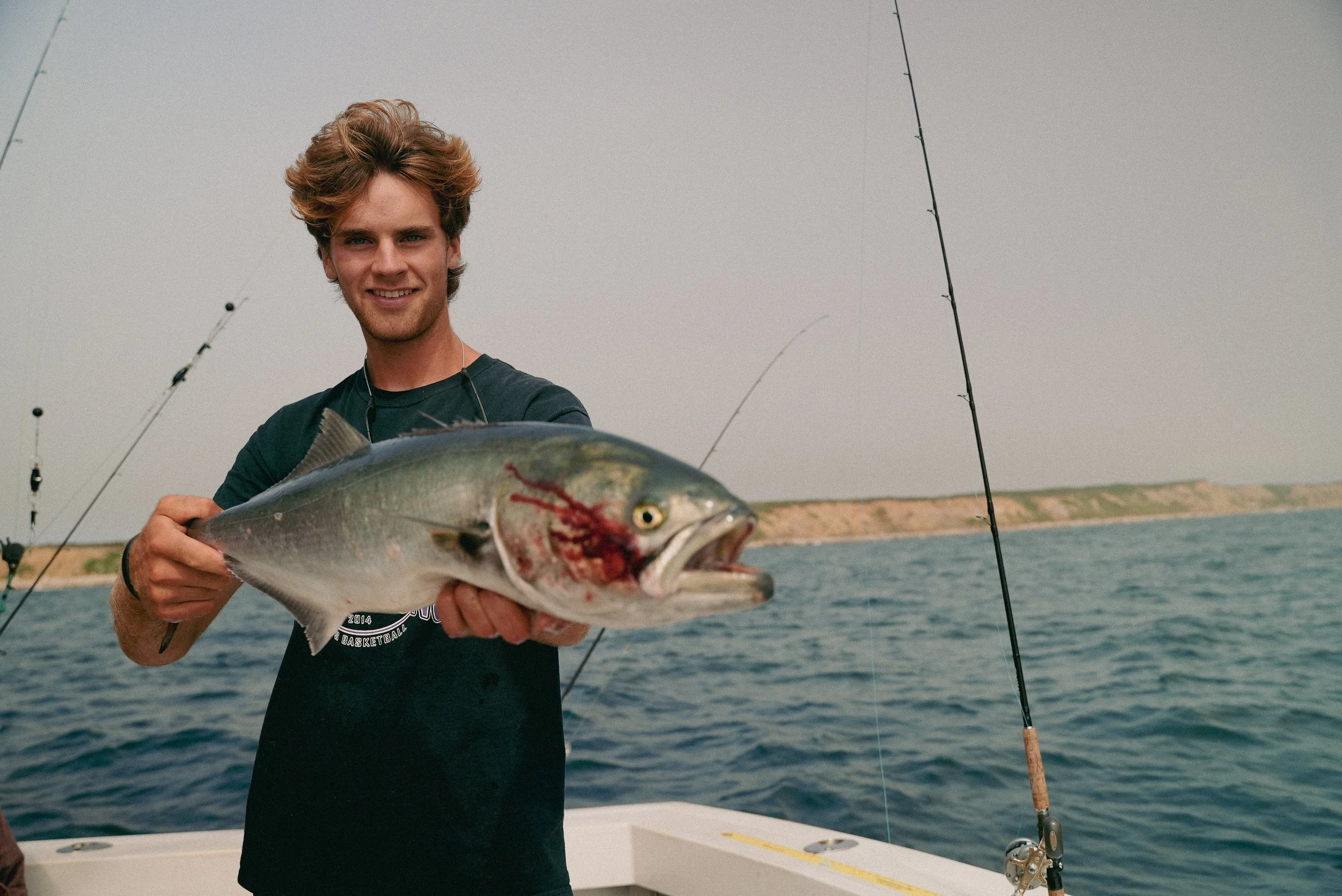 Person holding a fish on a boat with fishing rods and ocean backdrop.