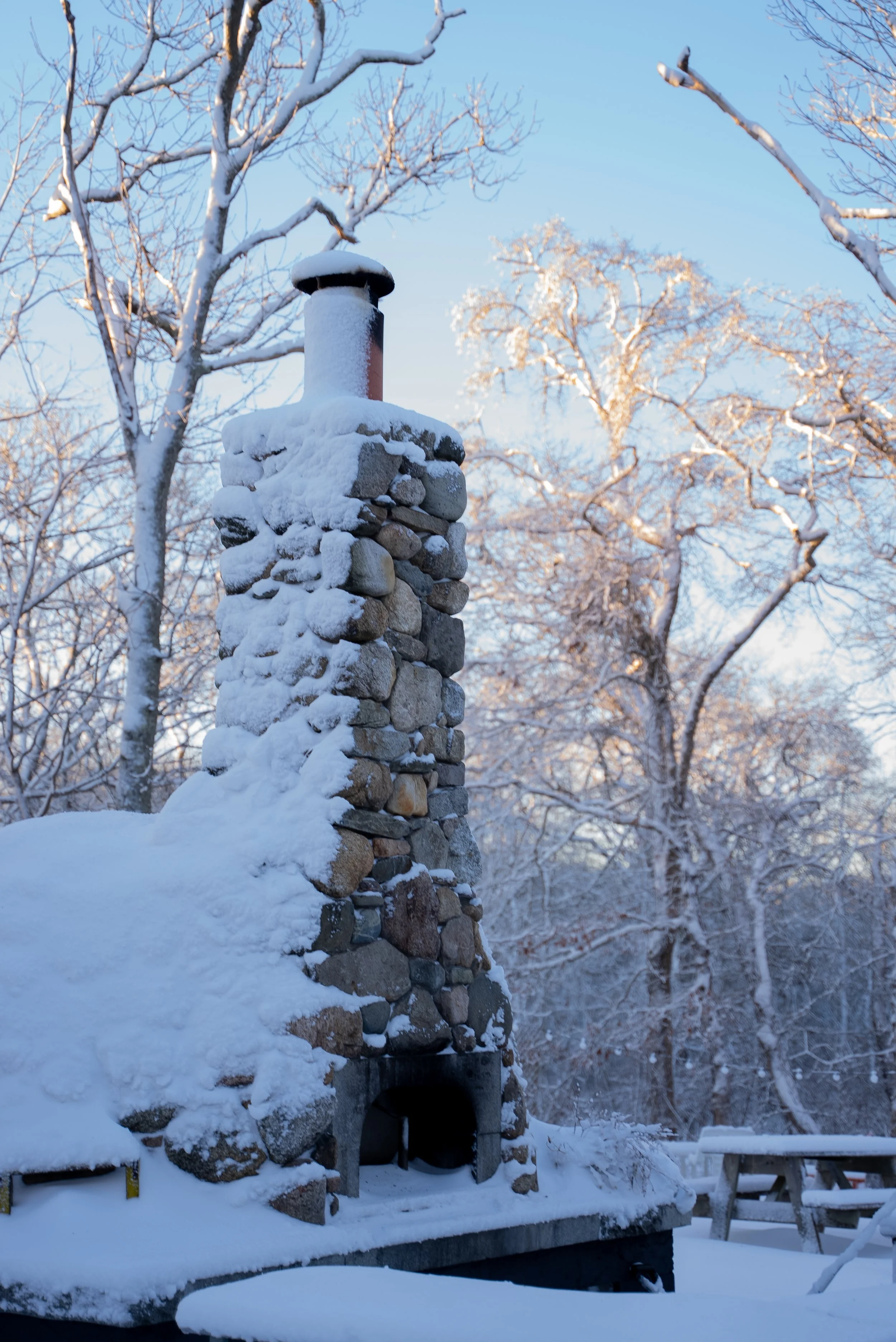 Stone chimney covered in snow with wintry trees and picnic bench in background.