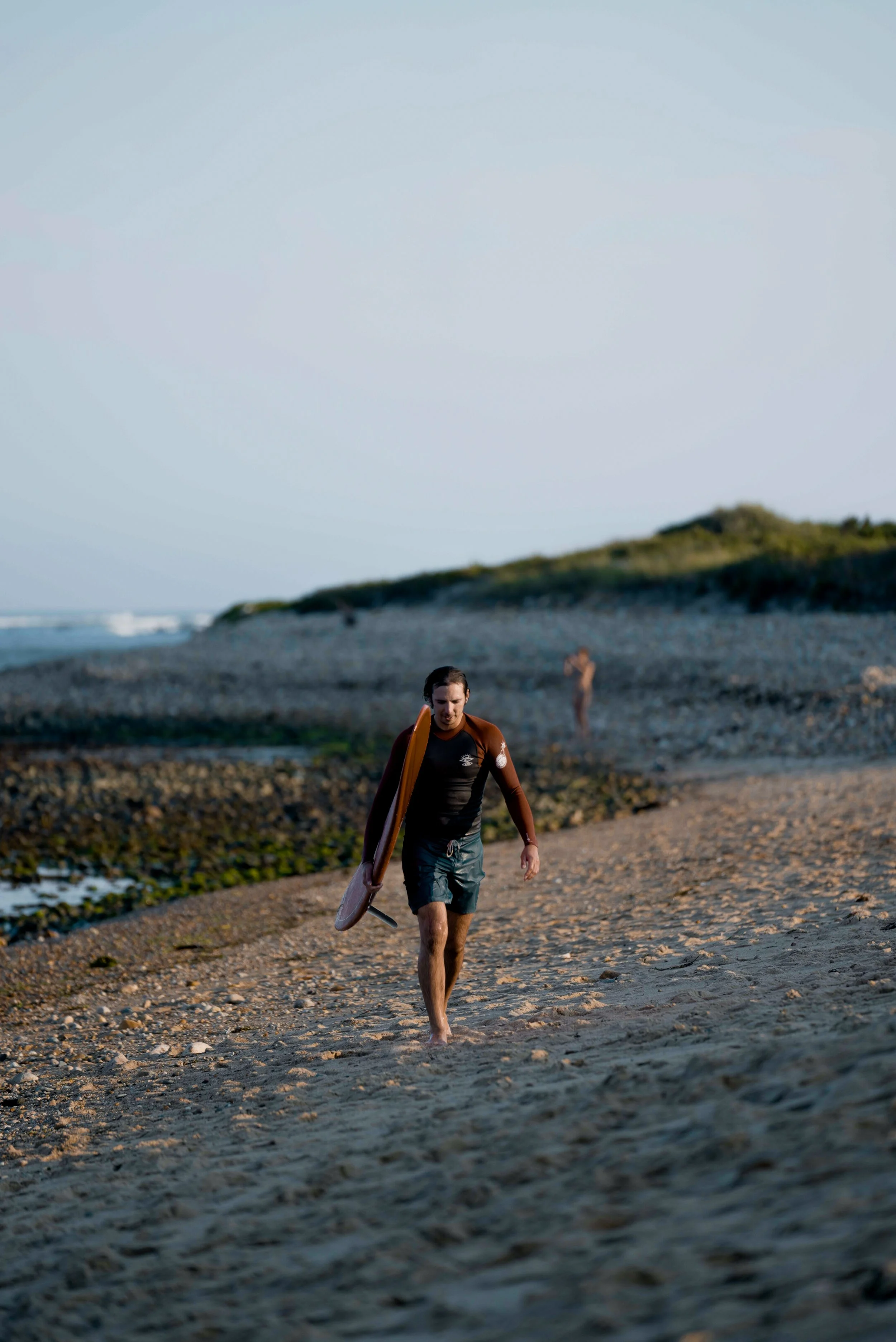 Surfer walking on a sandy beach with a surfboard at sunset.