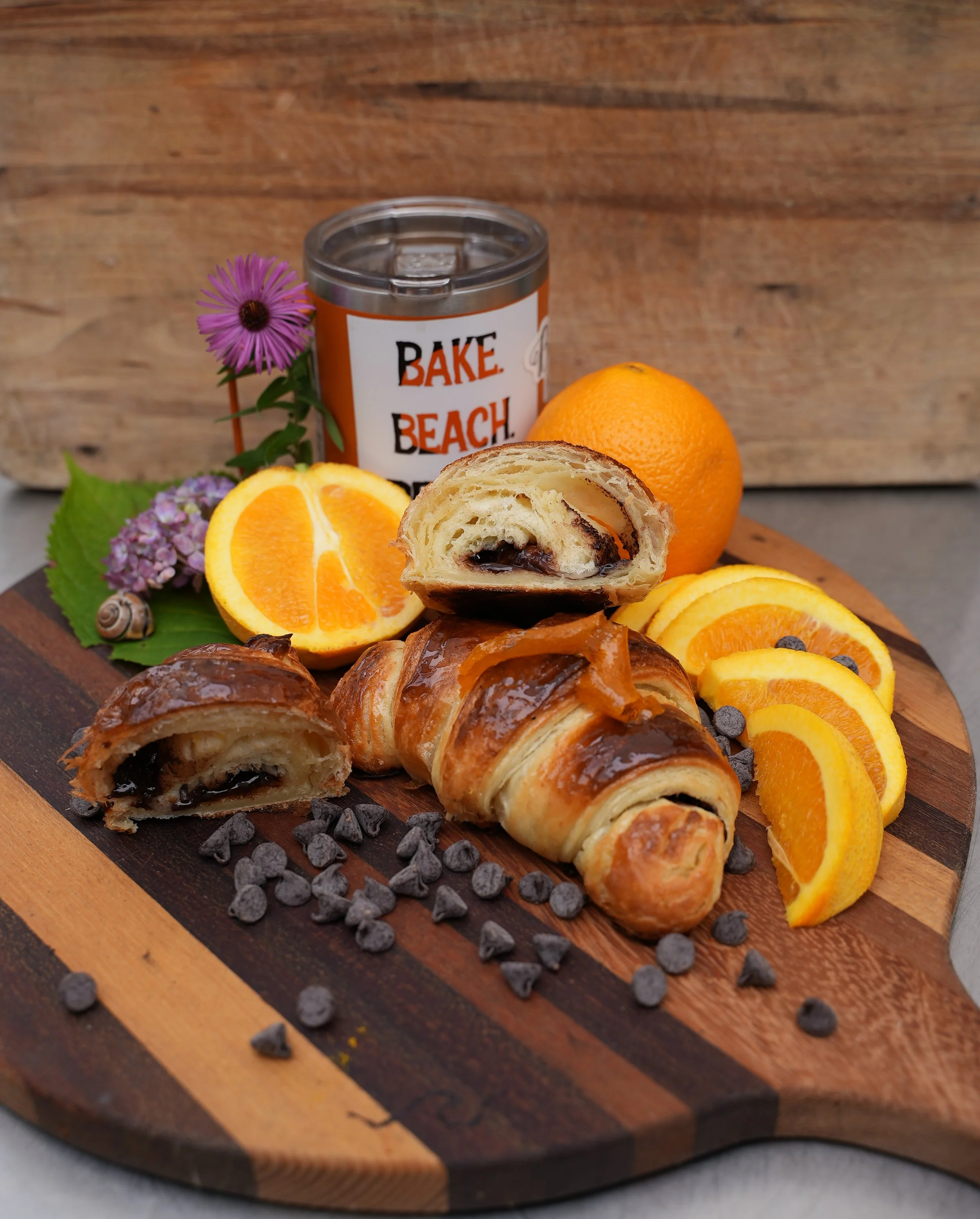 Croissants with chocolate chips and orange slices on a wooden board with a tin labeled "Bake Beach," flowers, and a snail shell.
