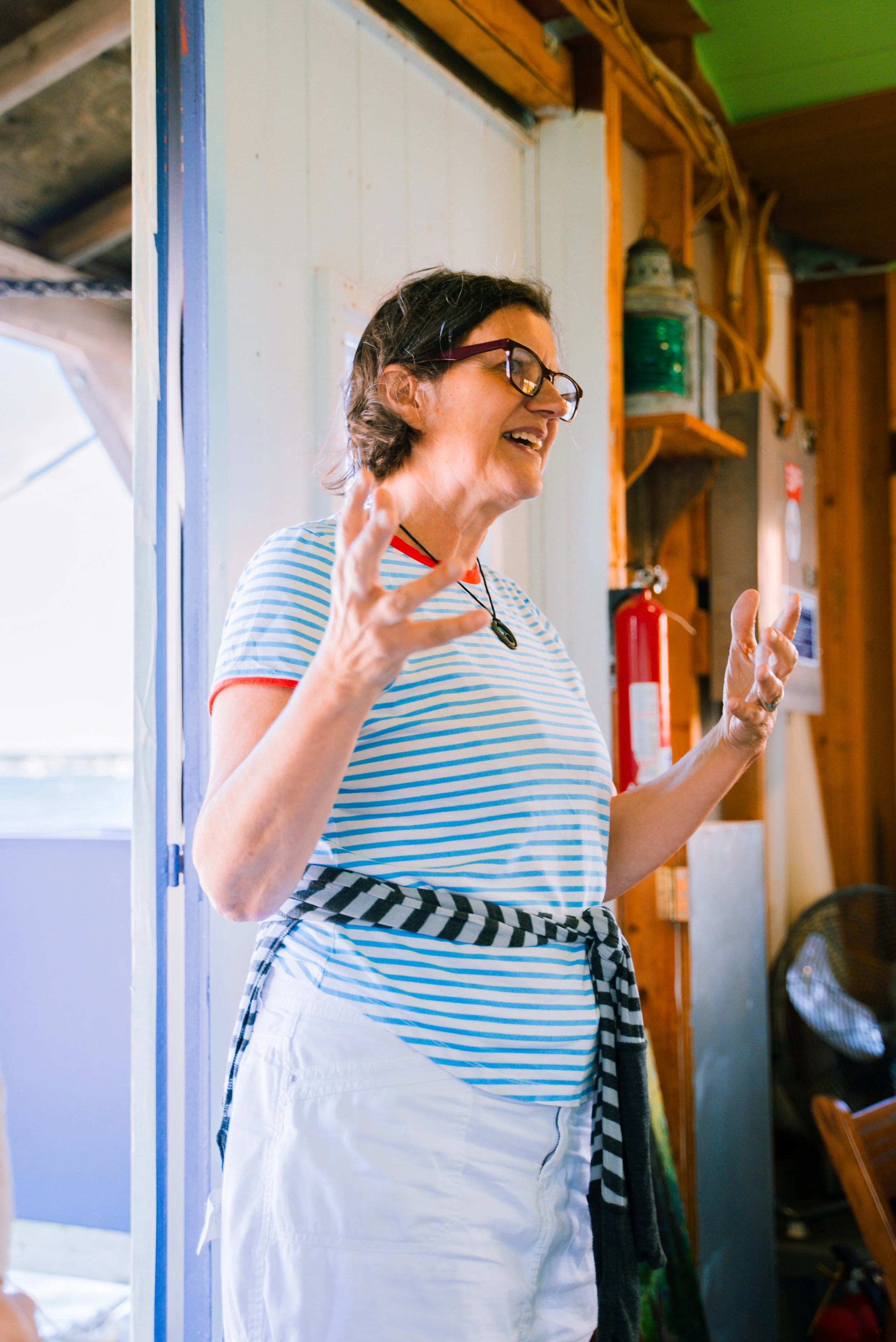 Person smiling, gesturing, wearing glasses and a striped shirt, indoors.