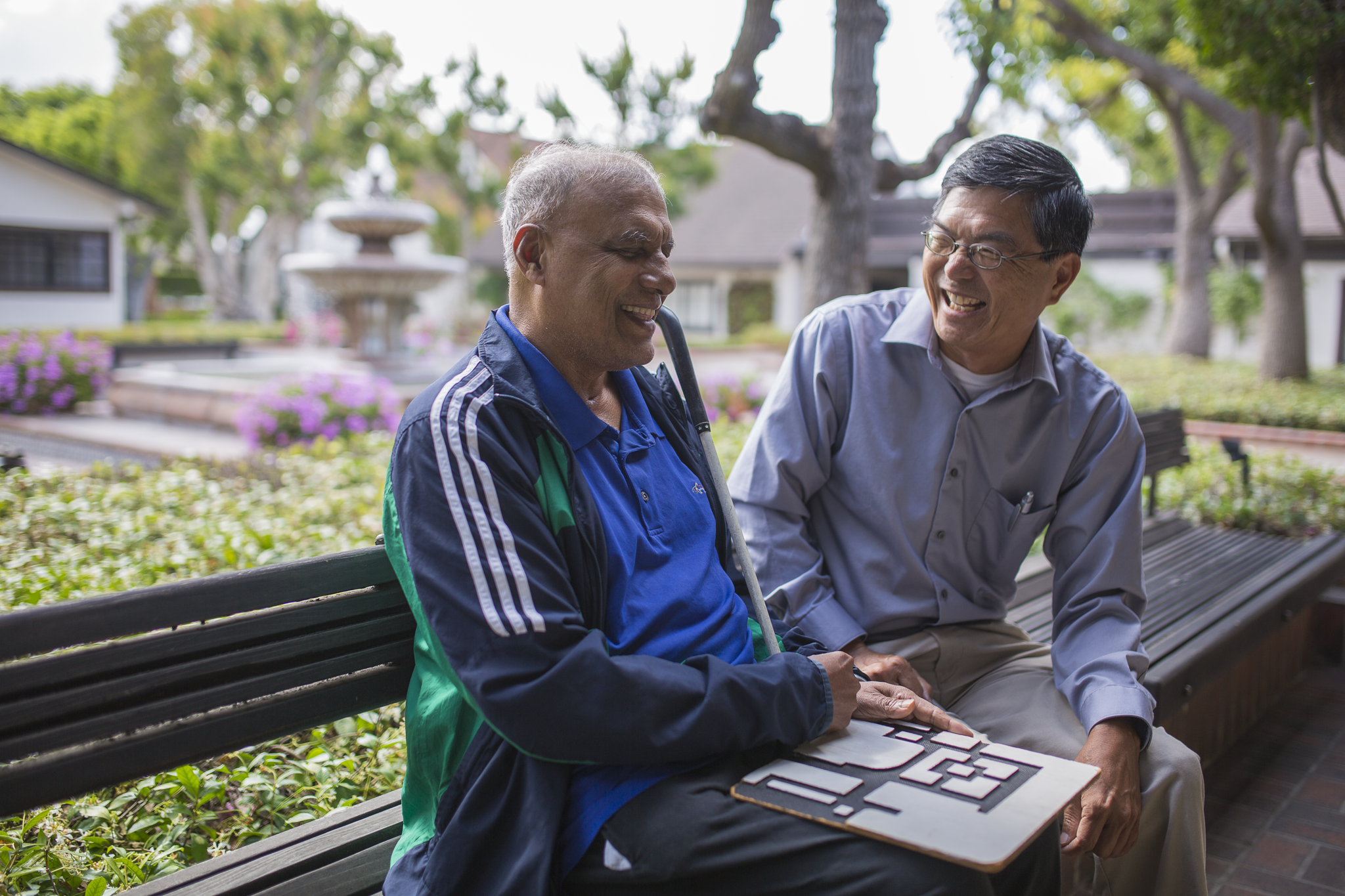 A man holding a white cane and a tactile map sitting on a bench laughing with another man wearing glasses.