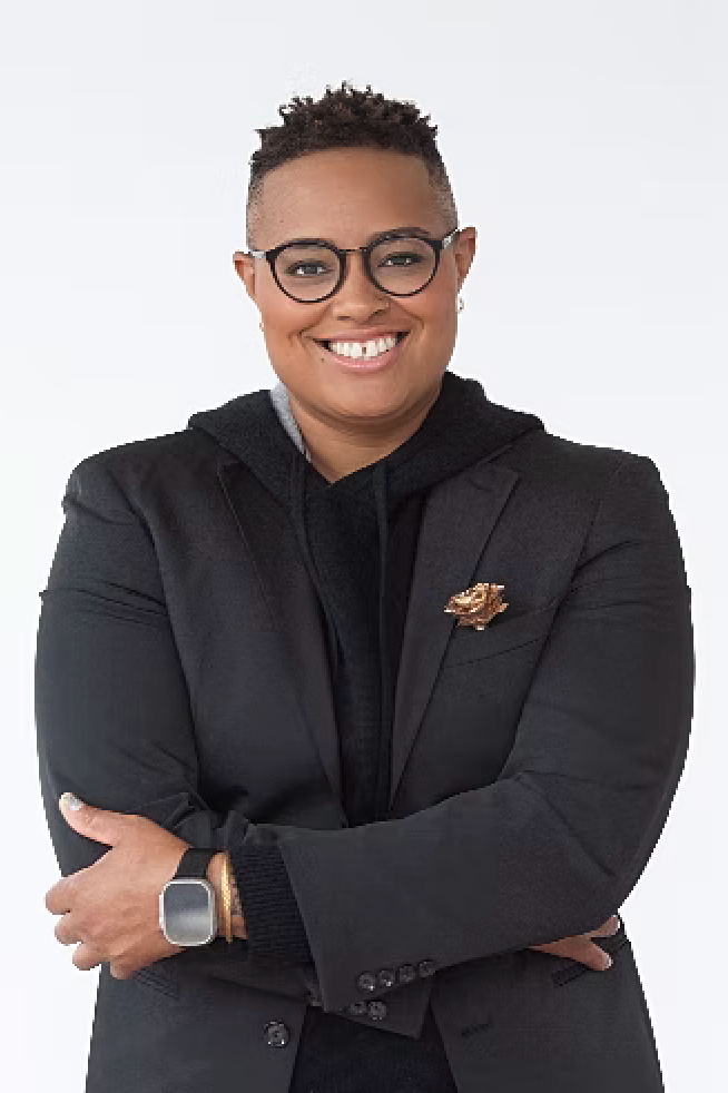 A smiling person with glasses, short curly hair, wearing a black blazer with a lion lapel pin, a black hoodie, a smartwatch, and crossing their arms against a white background.