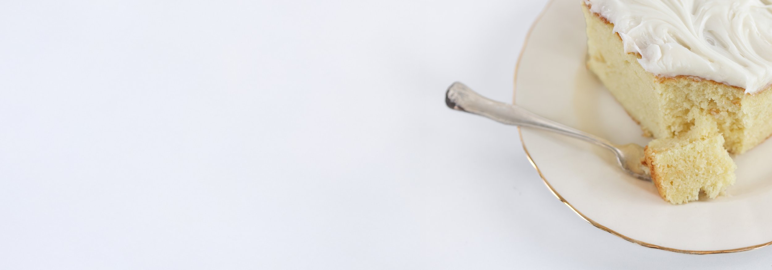 Slice of vanilla cake with white frosting and a blackberry on top, on a white plate with a small silver fork, set on a crumpled white cloth on a white surface.