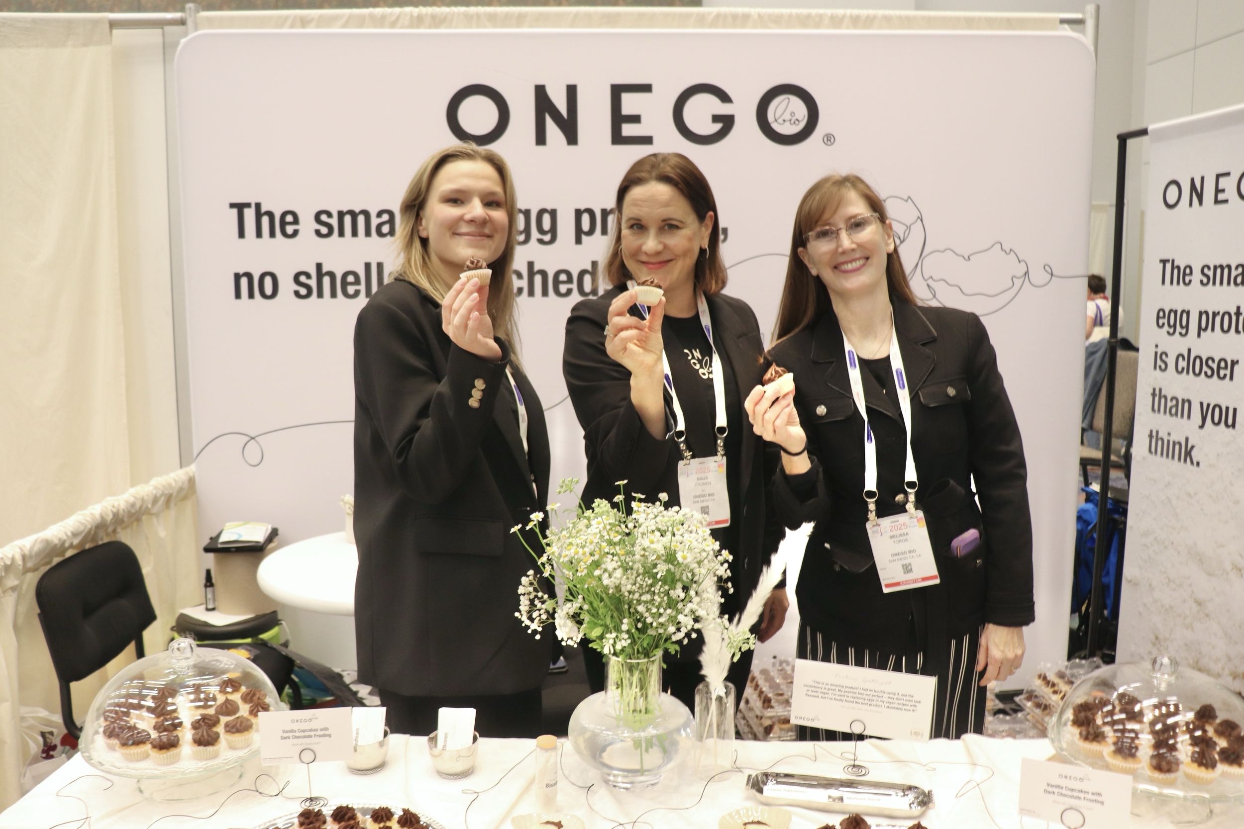 Three women standing behind a table with cupcakes, holding cupcakes and smiling. A large sign behind them reads 'ONEGO' and mentions egg-free products.
