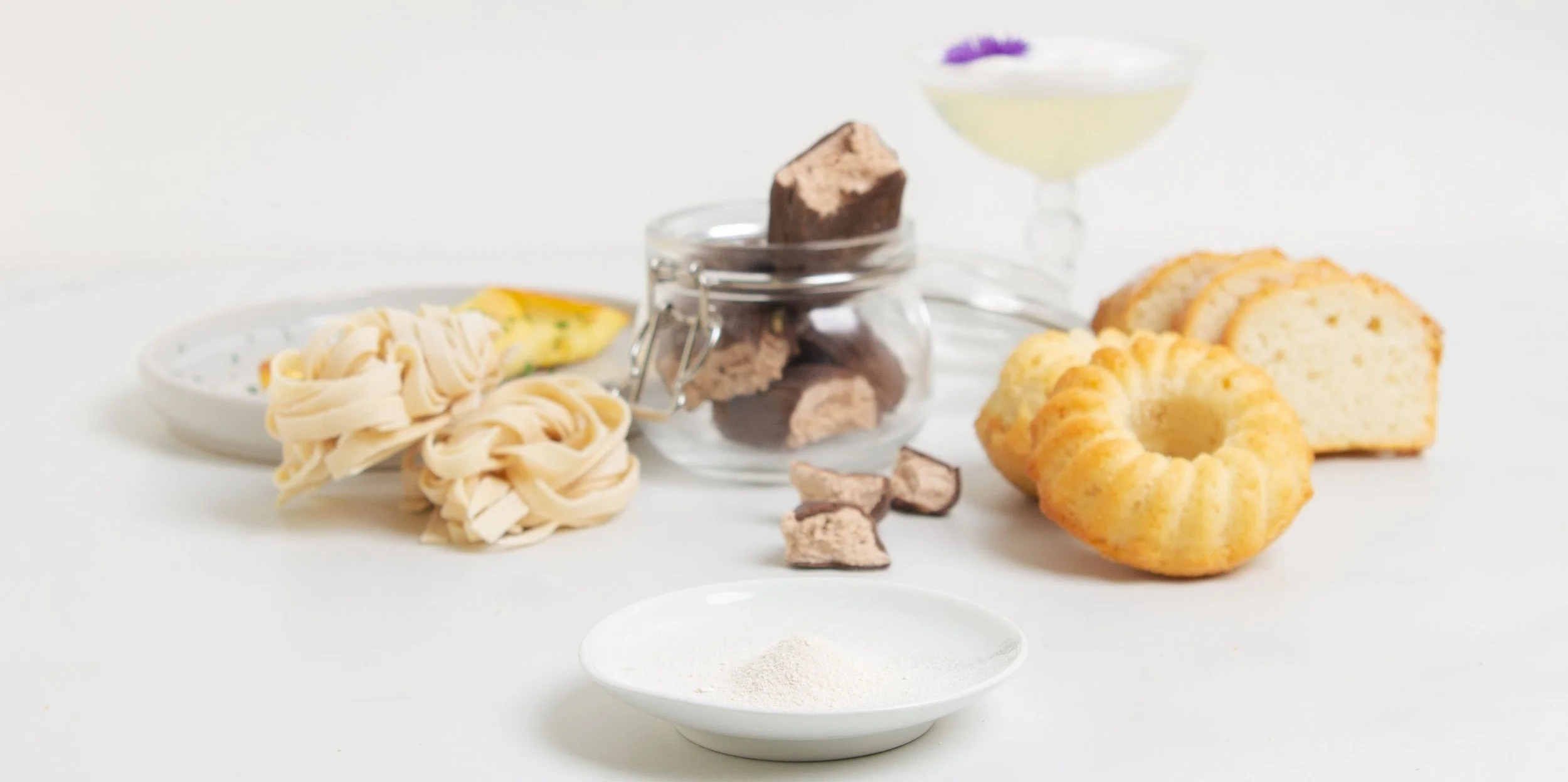 Various ingredients and baked goods on a white surface, including pasta, chocolate chunks, sliced pound cake, a bundt cake, and a cocktail with edible flowers.