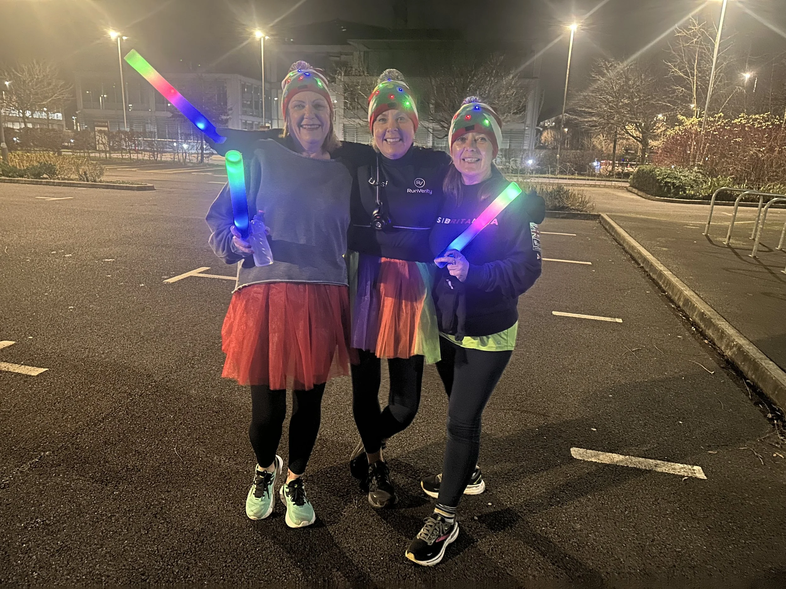 Three women in colorful attire holding glowing sticks, standing in a dimly lit parking lot at night.
