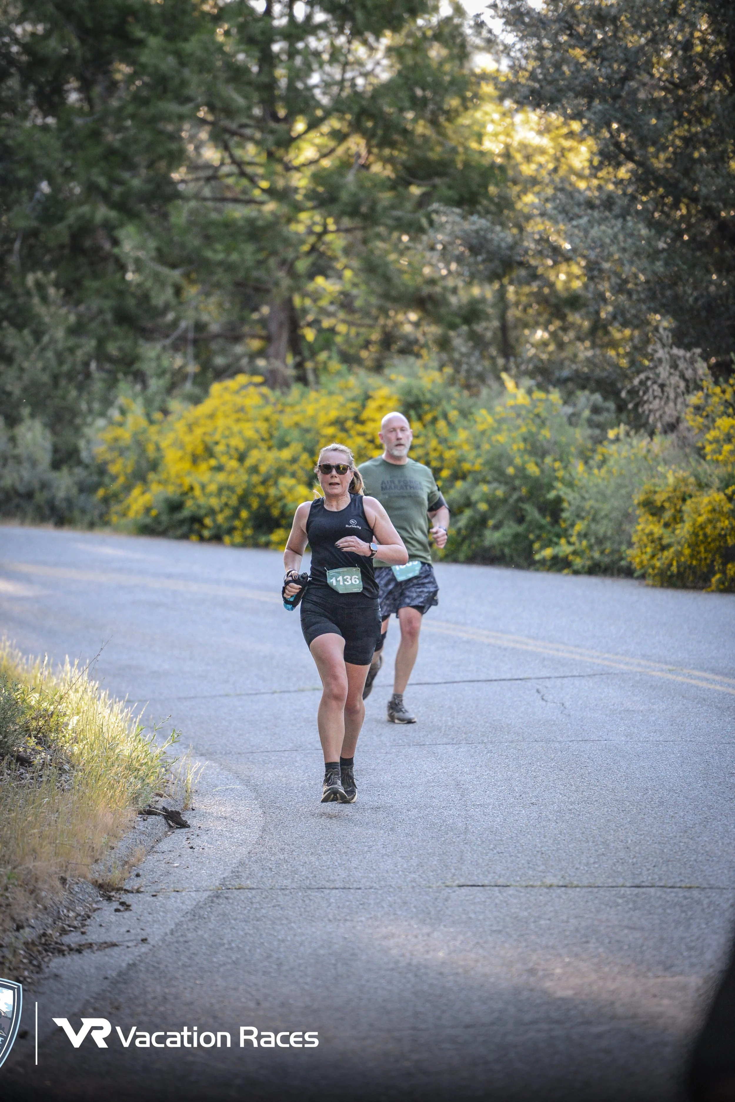 A woman and a man running on a paved road surrounded by trees and yellow flowering bushes during a VR Vacation Races marathon or race event.