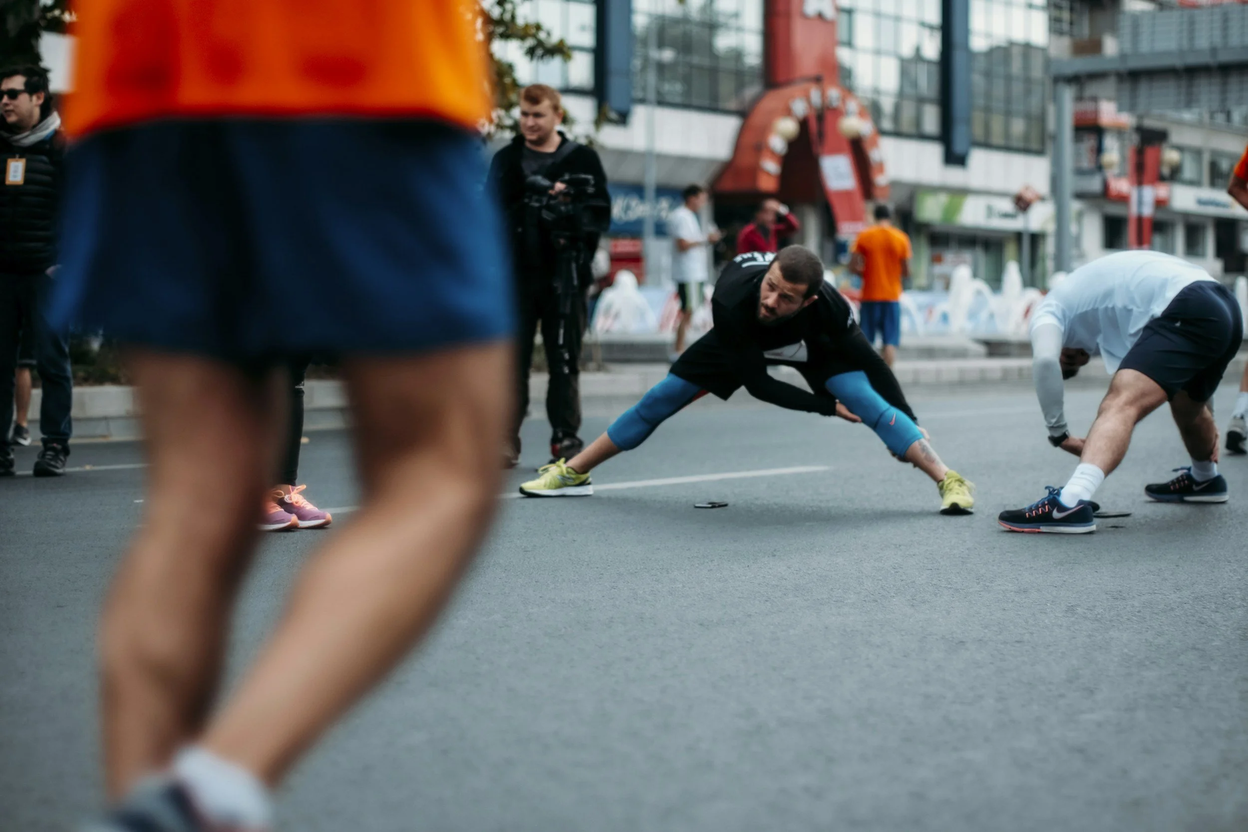 A group of people stretching and warming up on an urban street during a running or athletic event, with a man in black and blue stretching his leg in the center.
