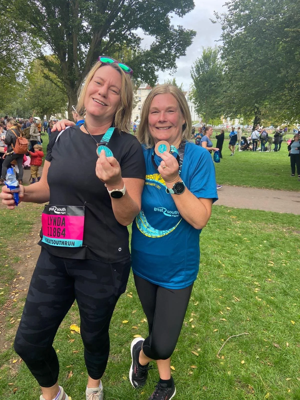 Two women holding medals after completing the Great South Run, standing in a park, wearing athletic attire, with a crowd and trees in the background.