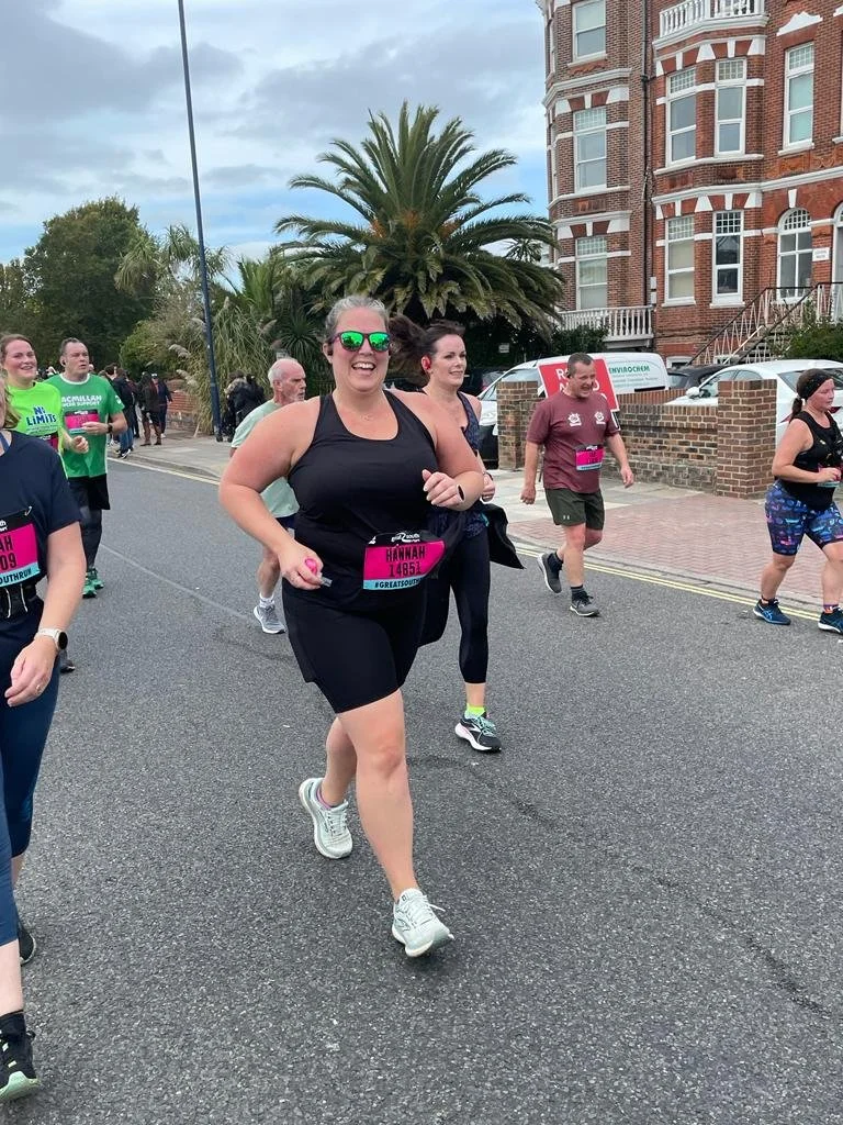 Participants running in a road race with bibs, smiling, surrounded by palm trees and a brick building.