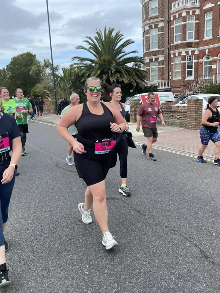 Group of people participating in a running event, smiling and jogging along a street lined with trees and a large brick building in the background.