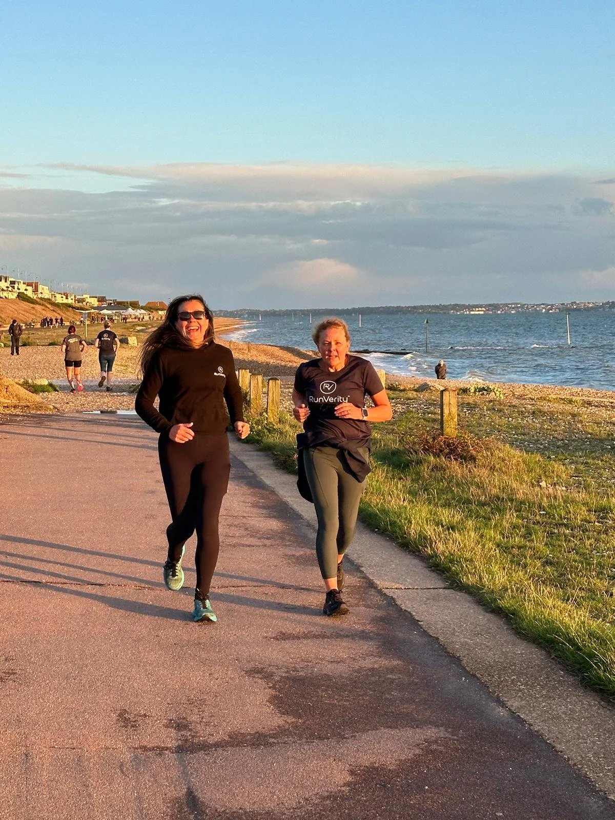 Two women jogging along a coastal path during sunset, with the ocean and a beach on the right and grass on the left. Other joggers and people are visible in the background.
