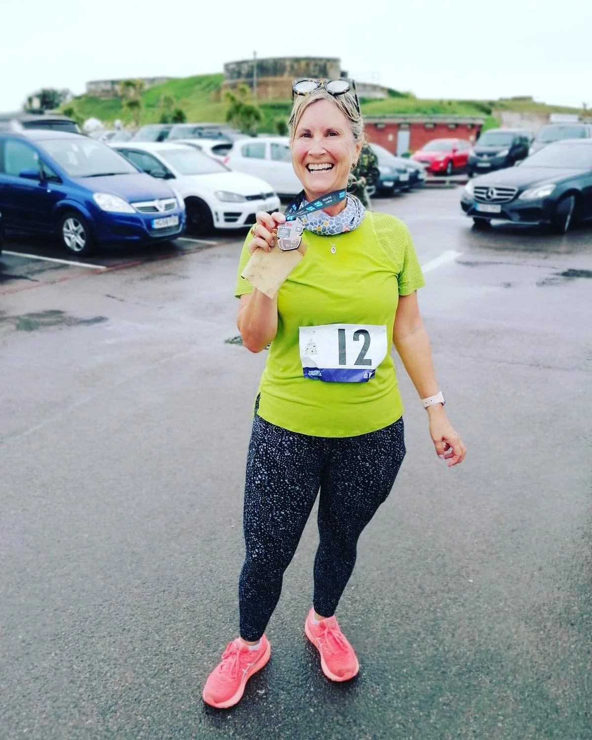 Smiling woman holding a medal, wearing a green shirt, patterned leggings, and pink sneakers in a parking lot.