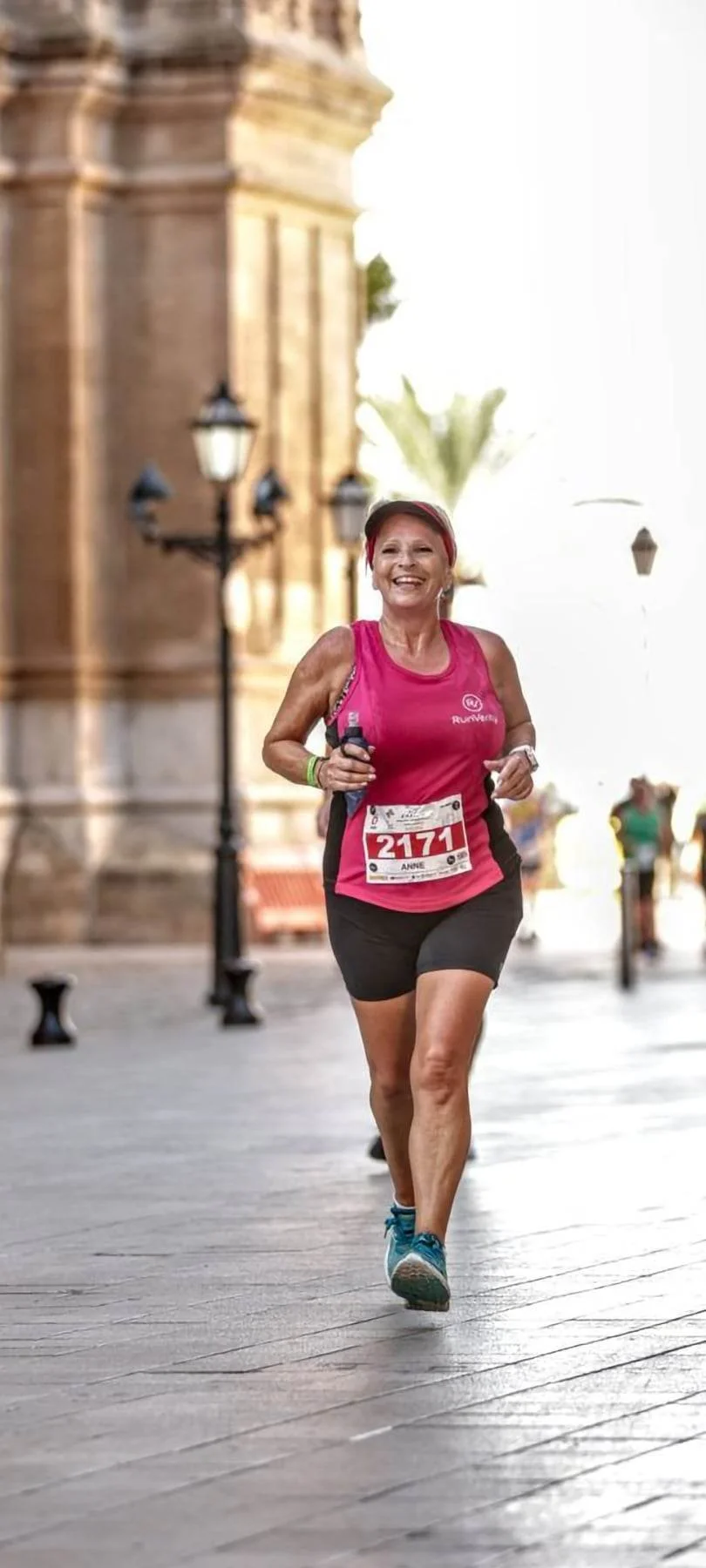 A woman running in a pink tank top and black shorts with a race bib, smiling, on a city street.