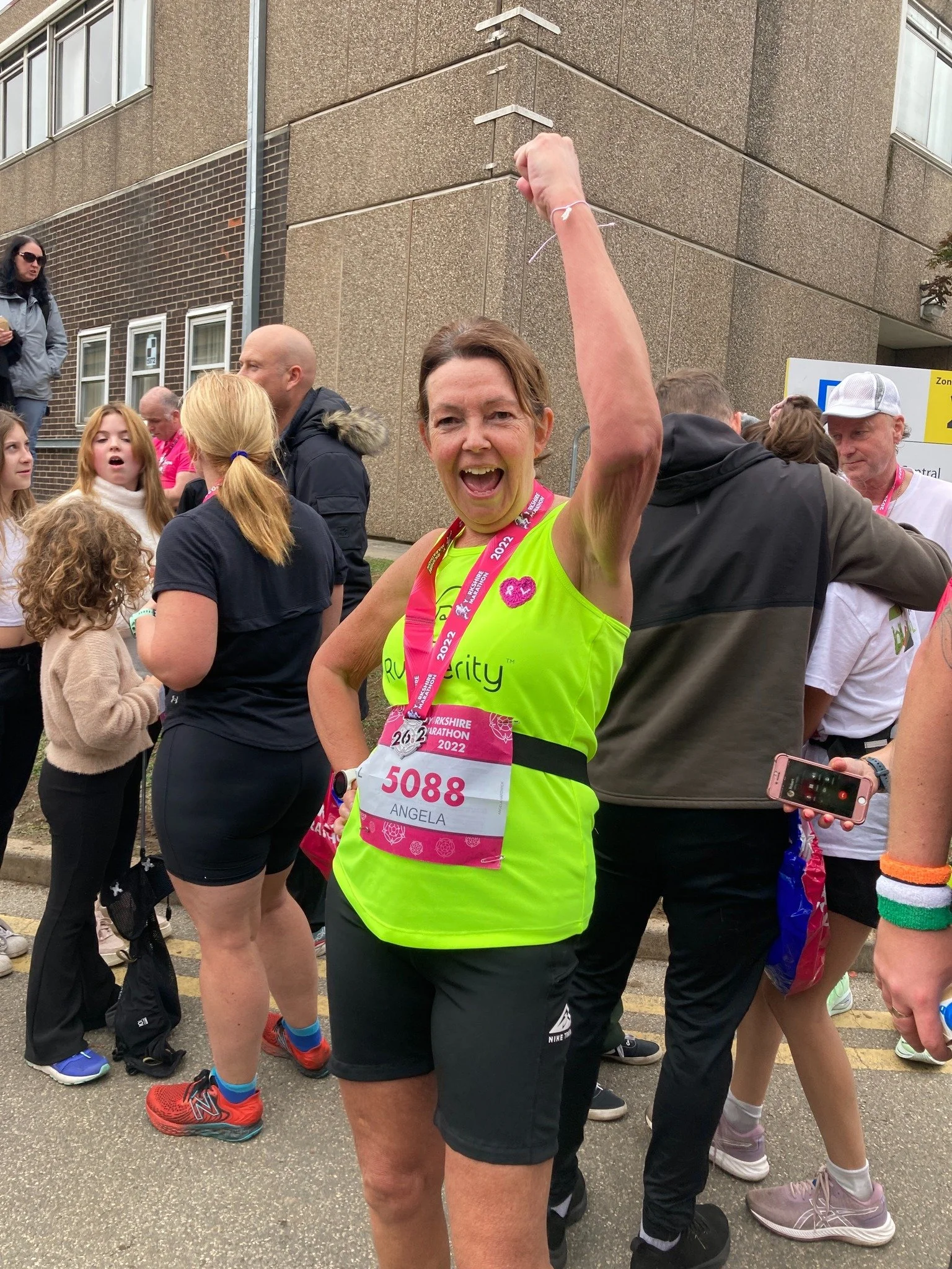 Woman celebrating after completing a marathon, wearing a bright green shirt and number 5088 bib, surrounded by other participants and bystanders.