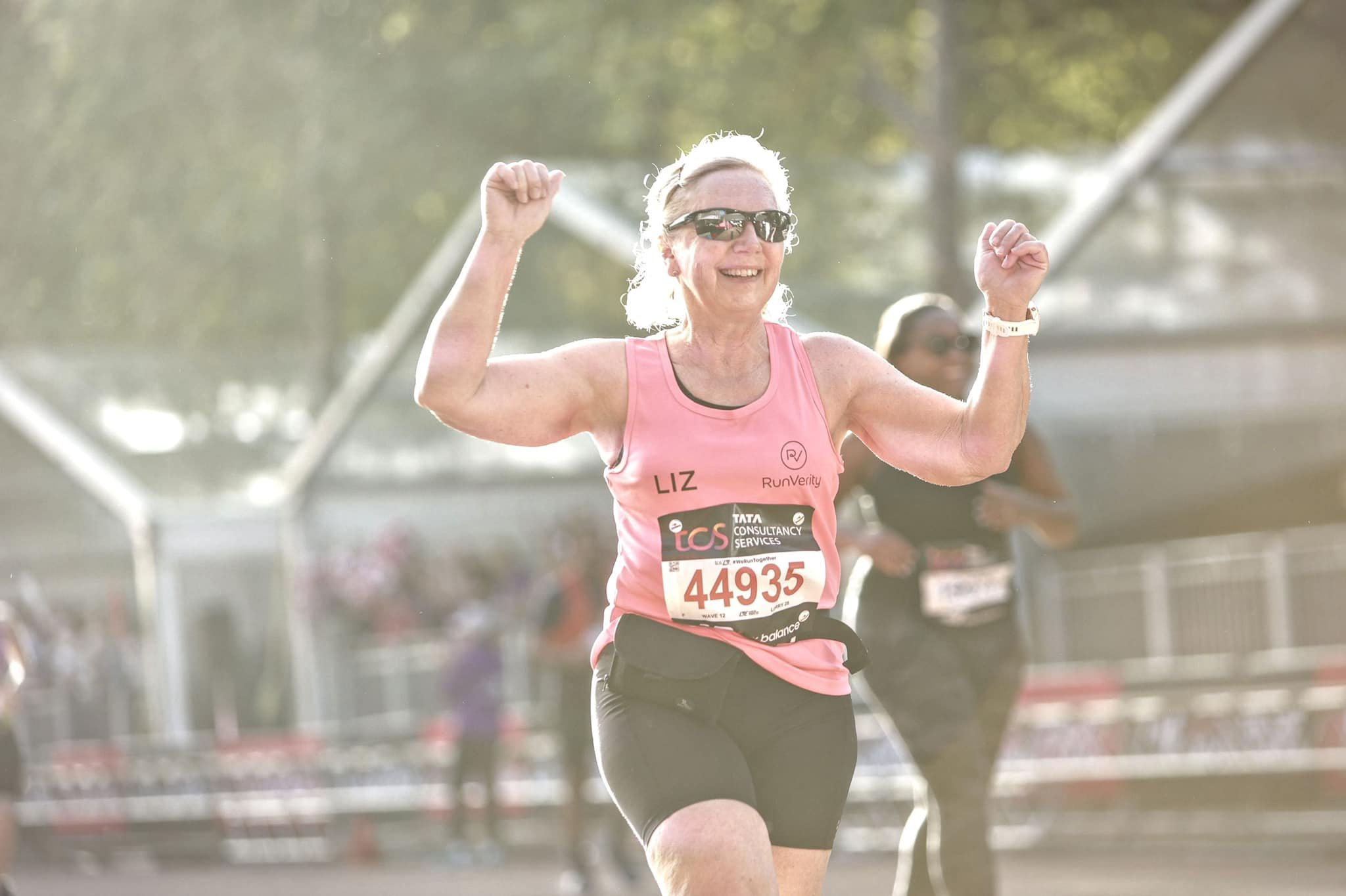 Woman with short hair running in a race wearing sunglasses, a pink tank top, and a race bib number 44935, arms raised in celebration.