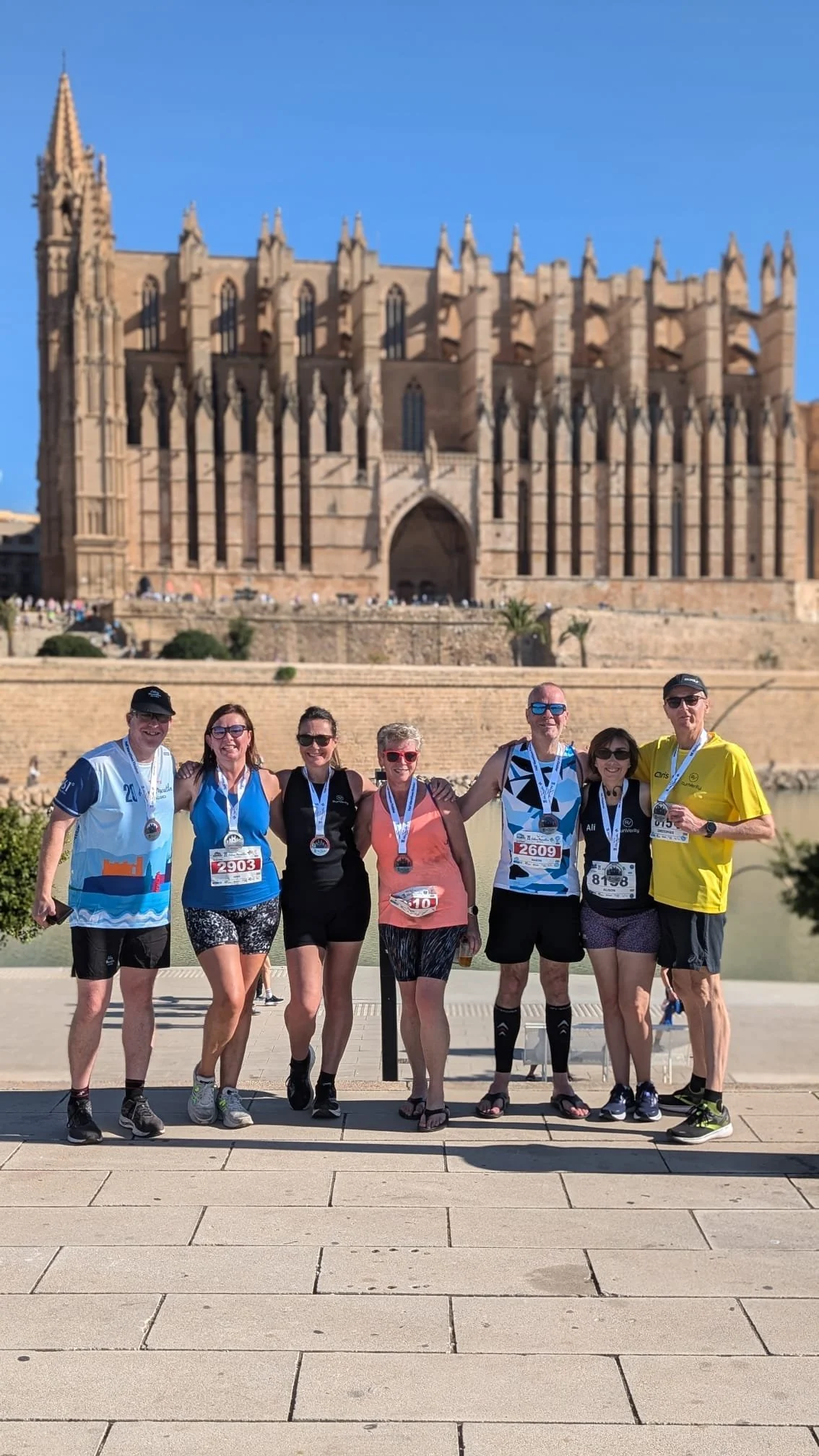 Seven runners wearing medals stand in front of a historic cathedral. They are dressed in sporty attire, indicating participation in a race. The cathedral features Gothic architecture with ornate spires and large arched windows.