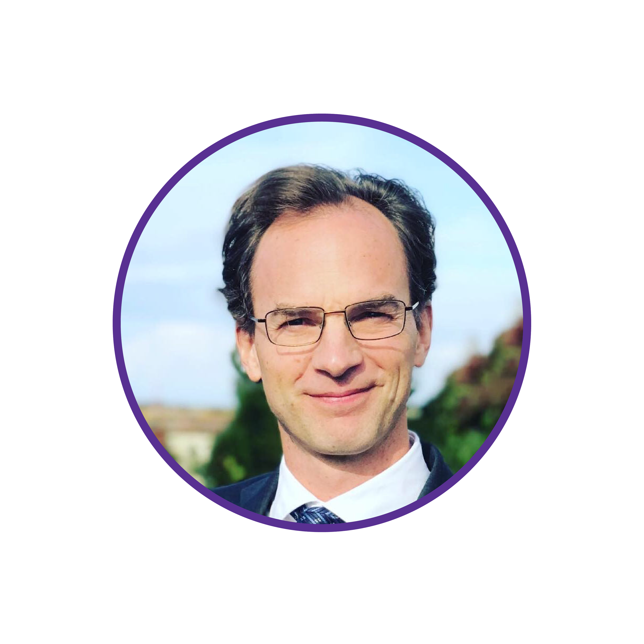 A man with glasses and dark hair smiling outdoors with trees and a cloudy sky in the background.