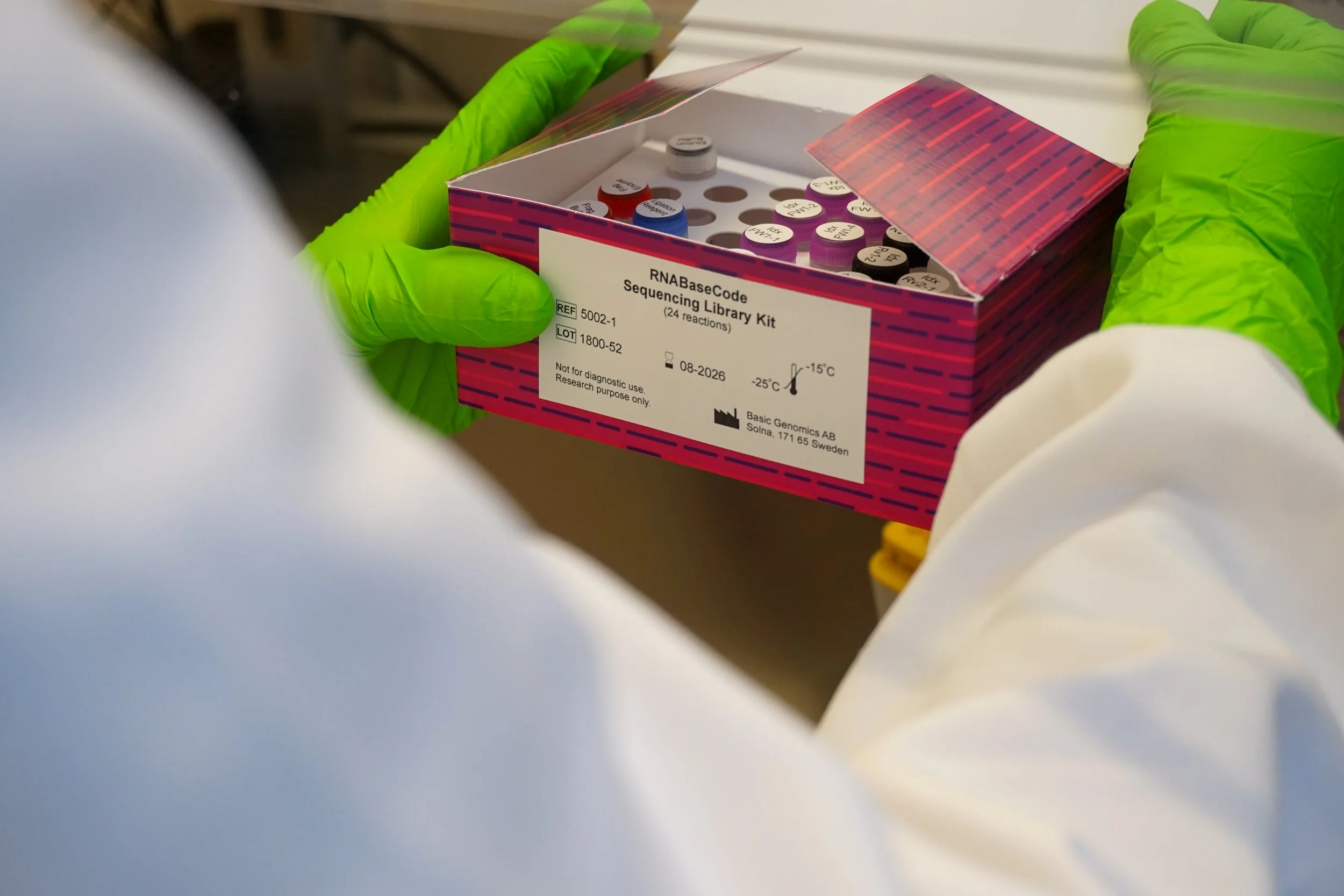 Person wearing green gloves holding a box labeled RNA BaseCode Sequencing Library Kit with samples inside