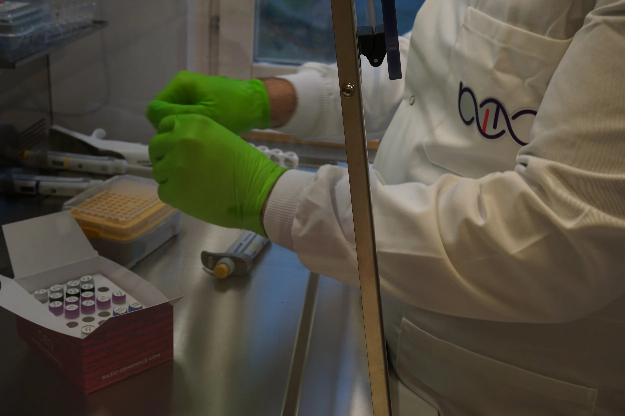 A scientist wearing a white lab coat and bright green gloves working with lab equipment inside a laboratory, handling test tubes or small vials.