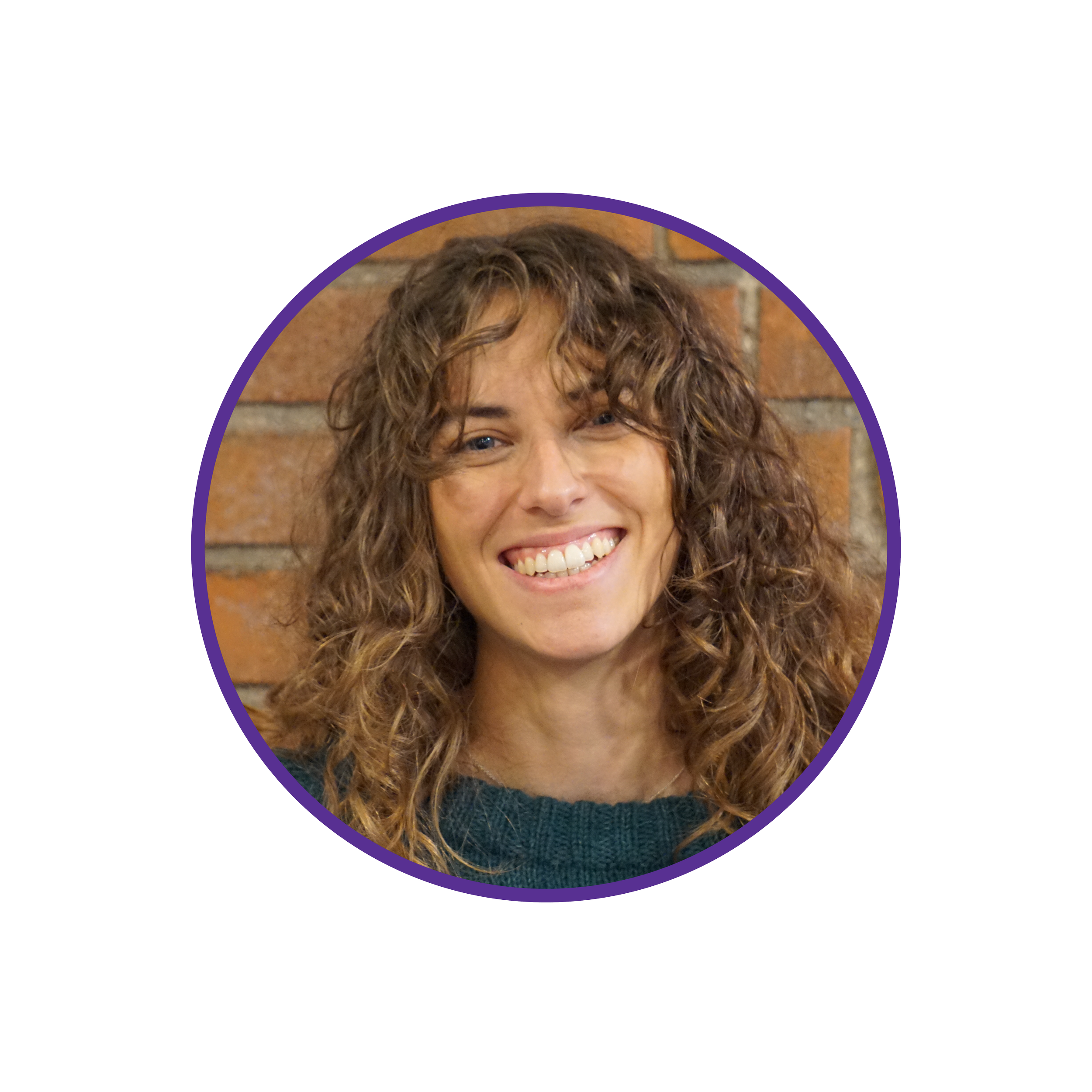 Smiling woman with curly brown hair, wearing a green sweater, standing in front of a brick wall.