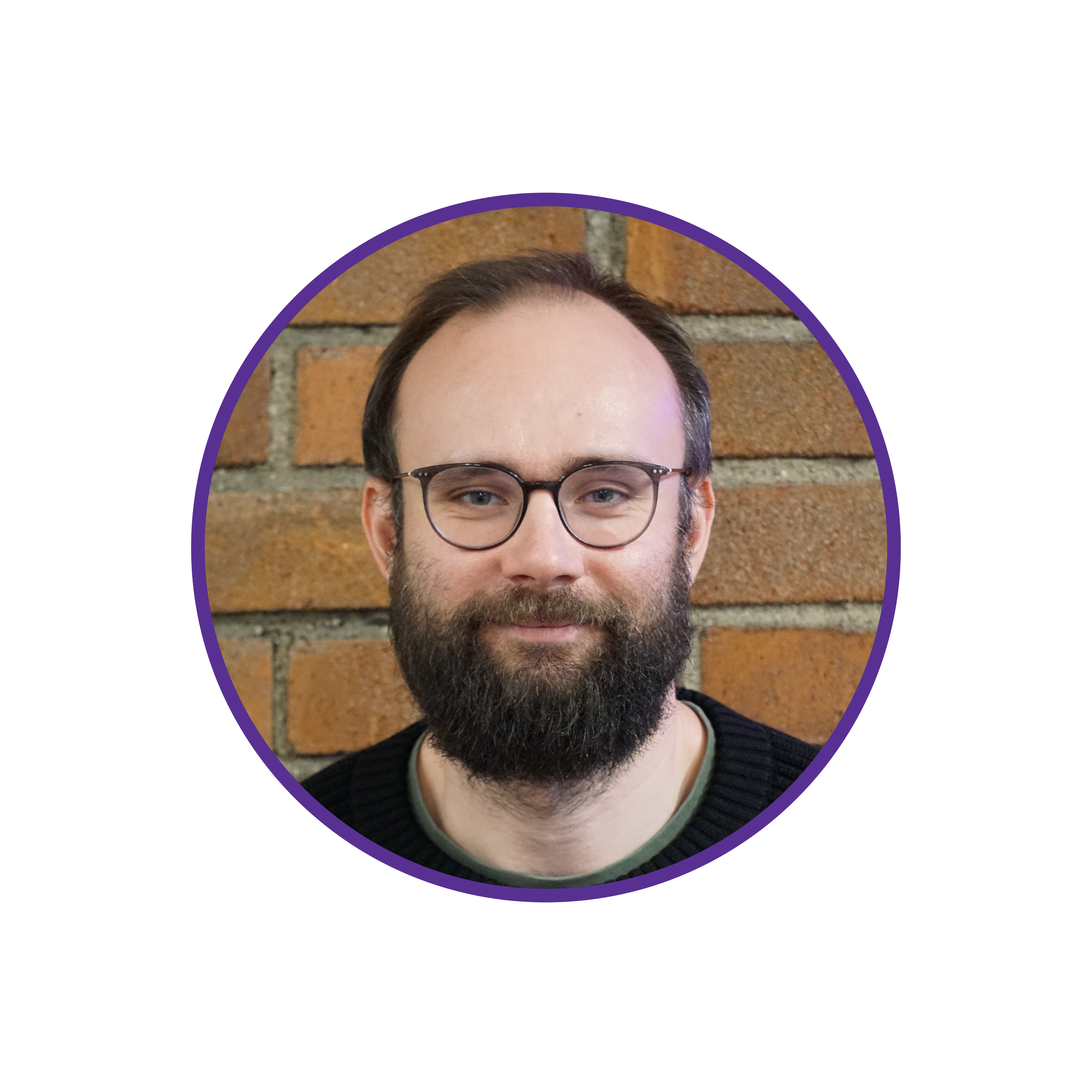 Headshot of a man with glasses and a beard, smiling, against a brick wall background.