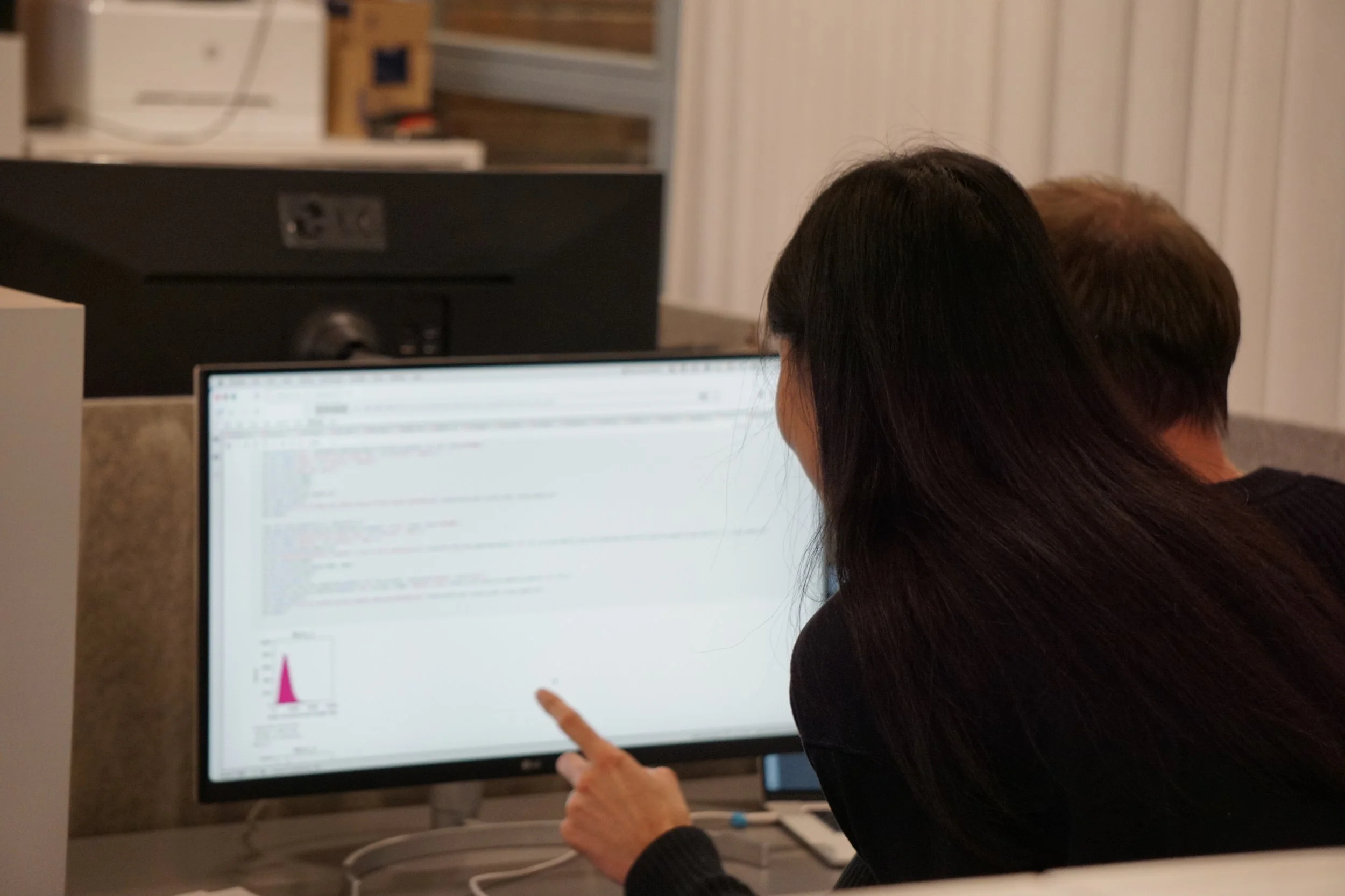 Two people working together on a computer in an office setting, with one person pointing at the monitor displaying code and graphs relating to RNA BaseCode data.