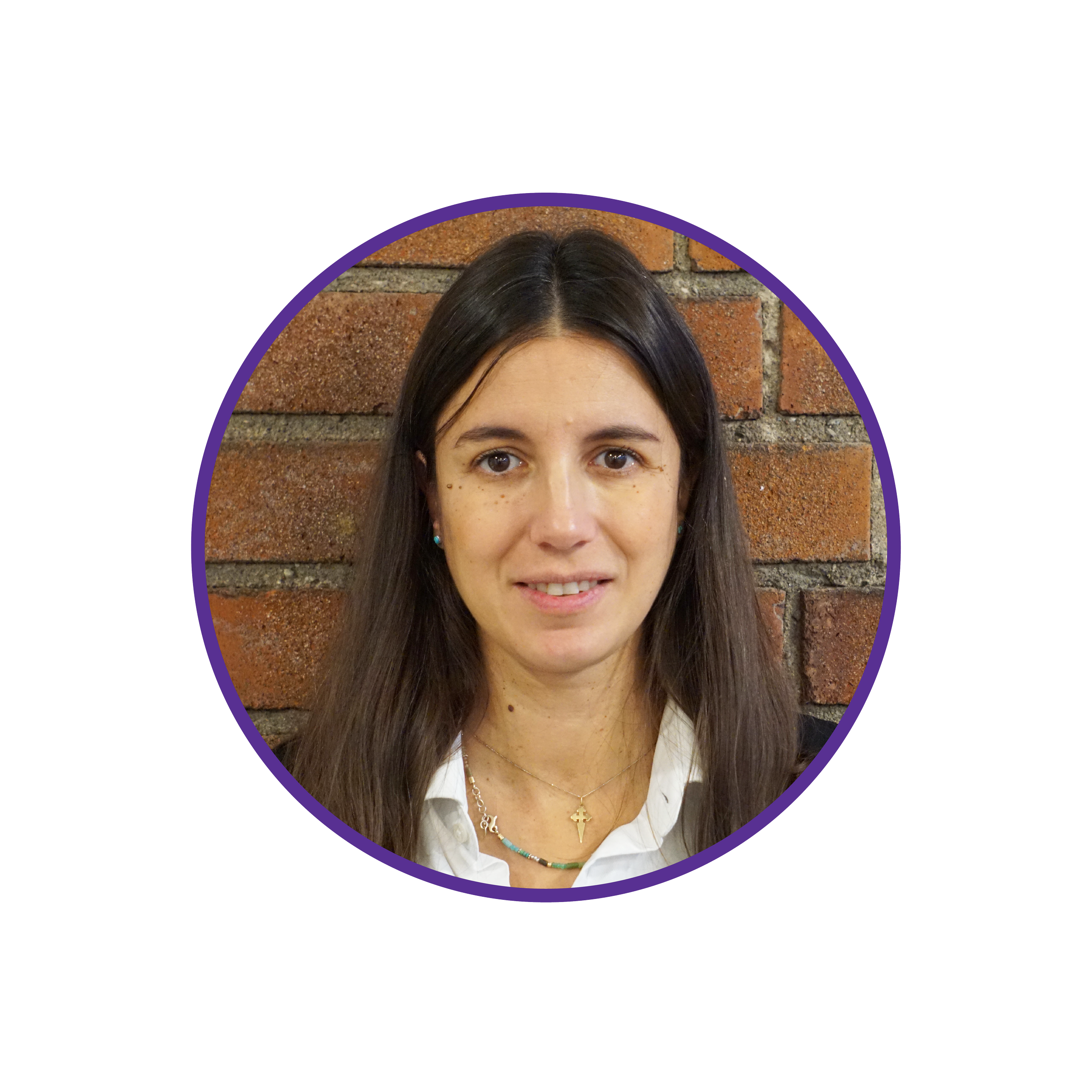 Headshot of a woman with long dark brown hair, light skin, standing against a brick wall, wearing a white shirt, gold necklace, and small earrings.