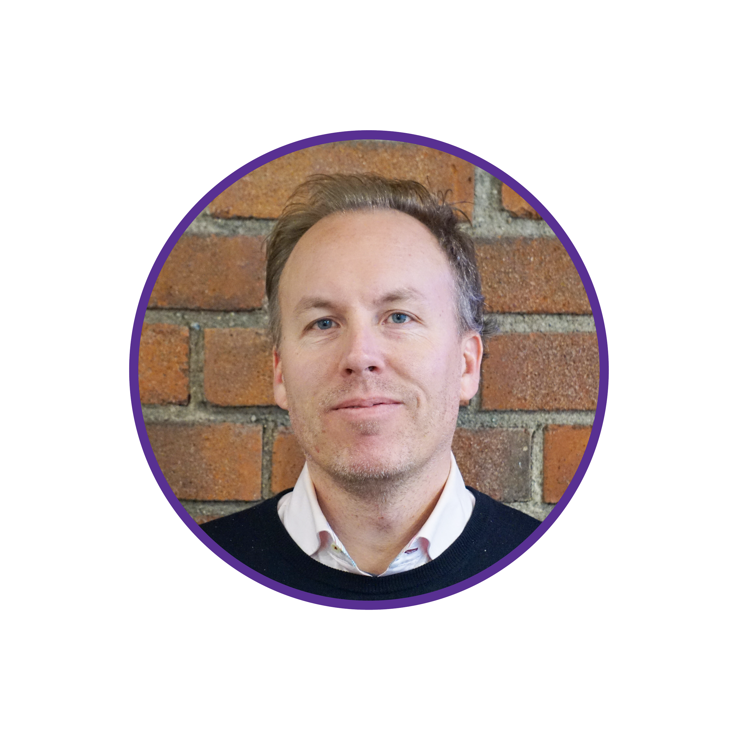 Headshot of a man with light brown hair, blue eyes, wearing a white collared shirt and a black sweater, posing in front of a brick wall.