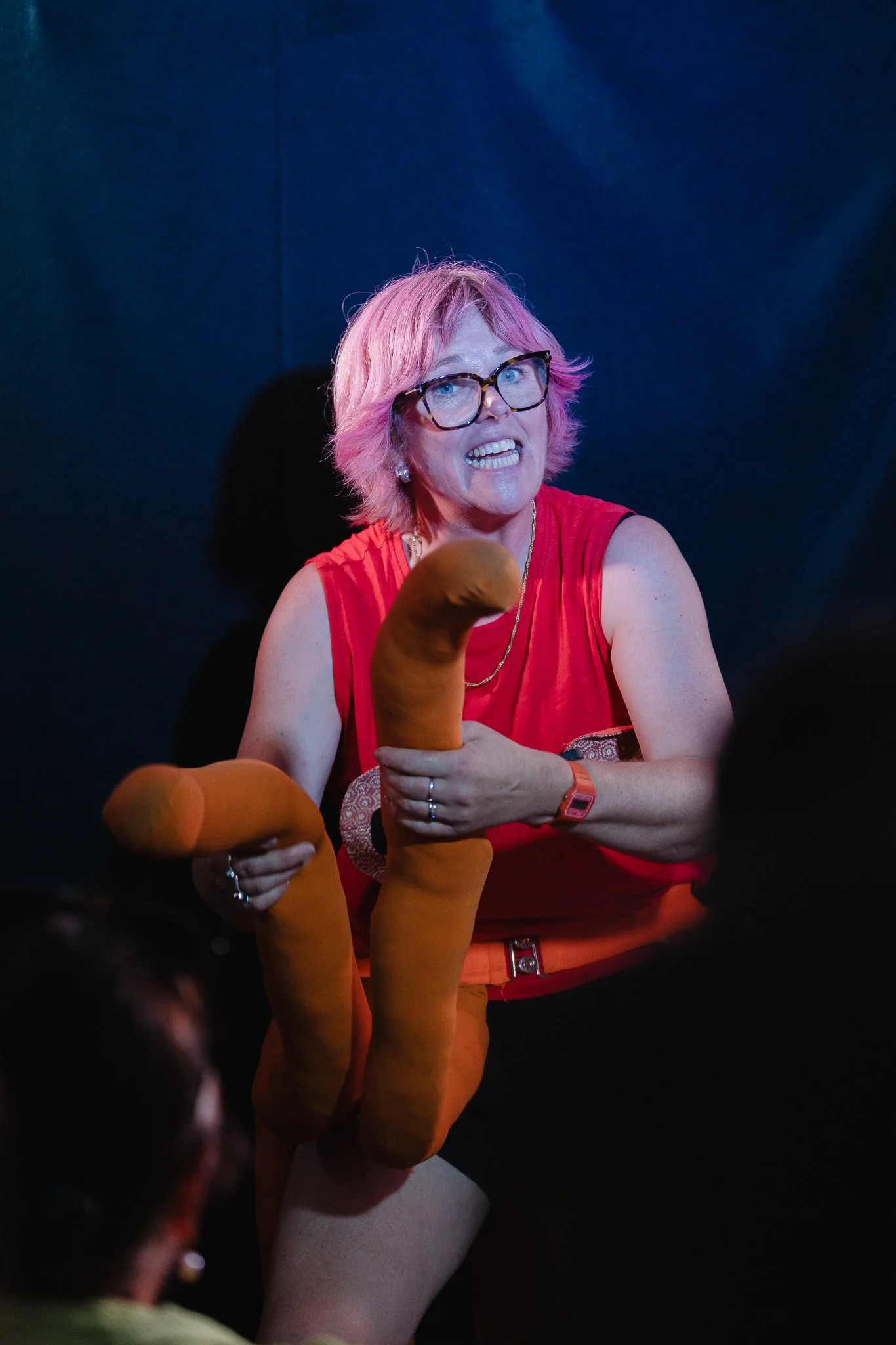 A woman with pink hair, glasses, and a red sleeveless top appears to be in an energetic setting, holding orange sock puppets resembling animals, possibly dogs, with a blue and dark background.