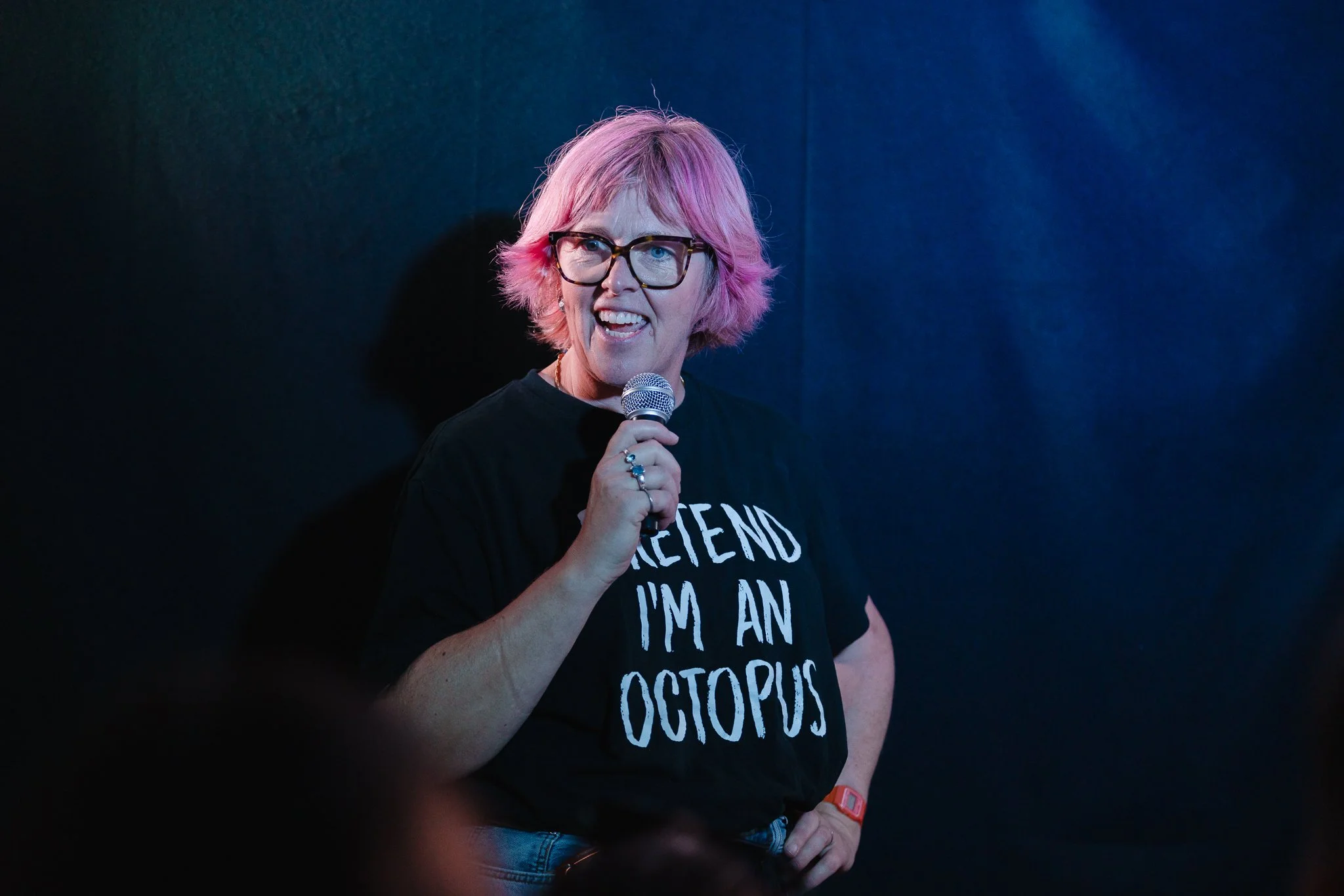 A woman with pink hair and glasses speaking into a microphone, wearing a black T-shirt with white text.