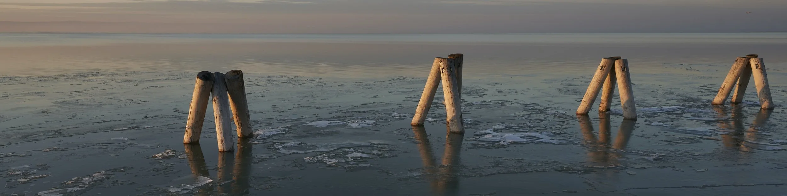 Holzpfeiler im gefrorenen See bei Sonnenuntergang, mit reflektierendem Wasser und Eisflächen.