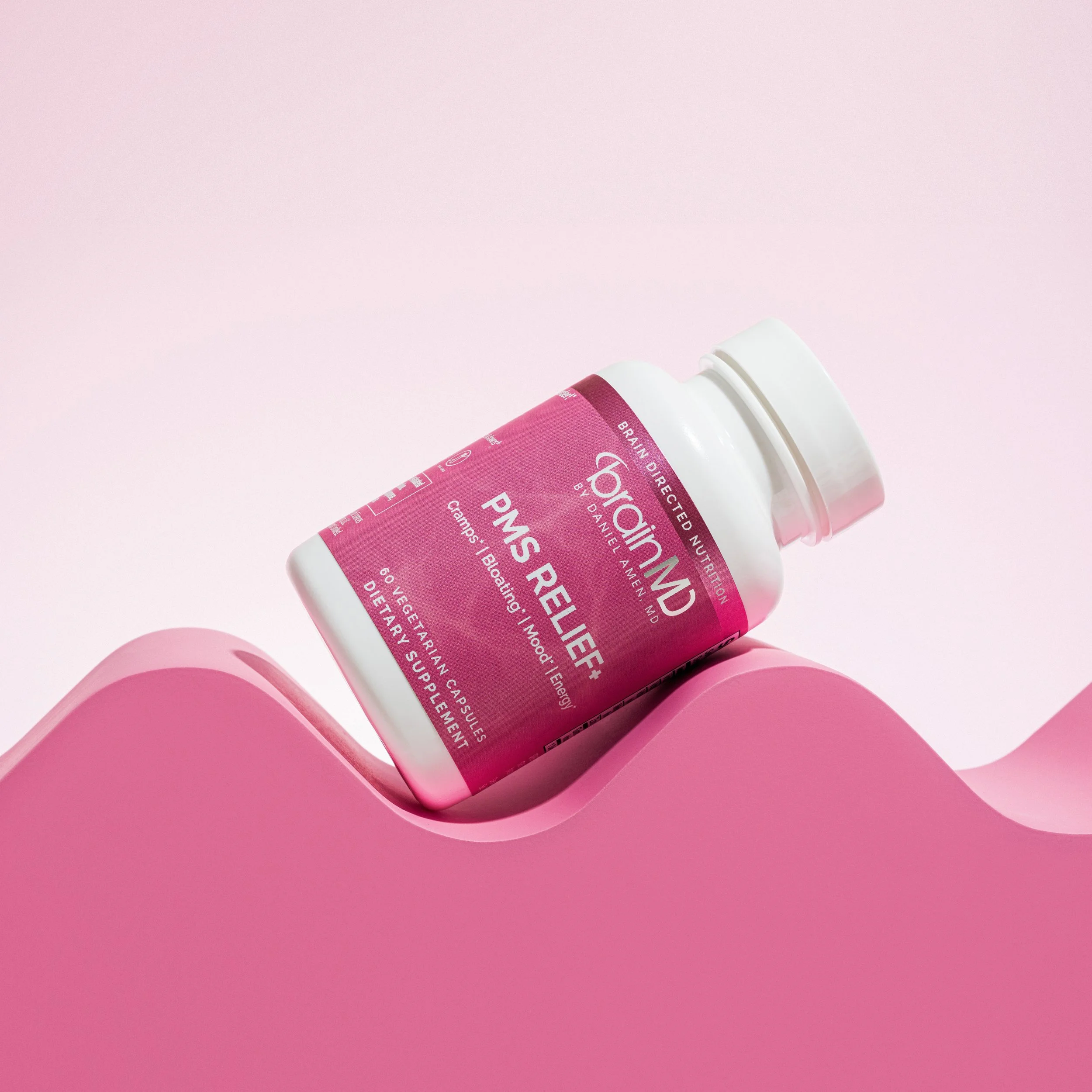 A pink bottle labeled 'BrainMD PNS Relief' lying on a pink surface with a curved design, against a light pink background.