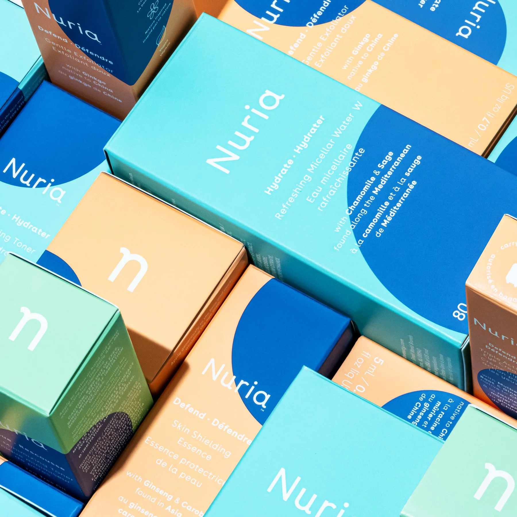 Multiple boxes of a skincare product labeled Nuria in blue, peach, and green packaging with descriptive text about hydrating and defending skin, with key ingredients like ginseng and chamomile.