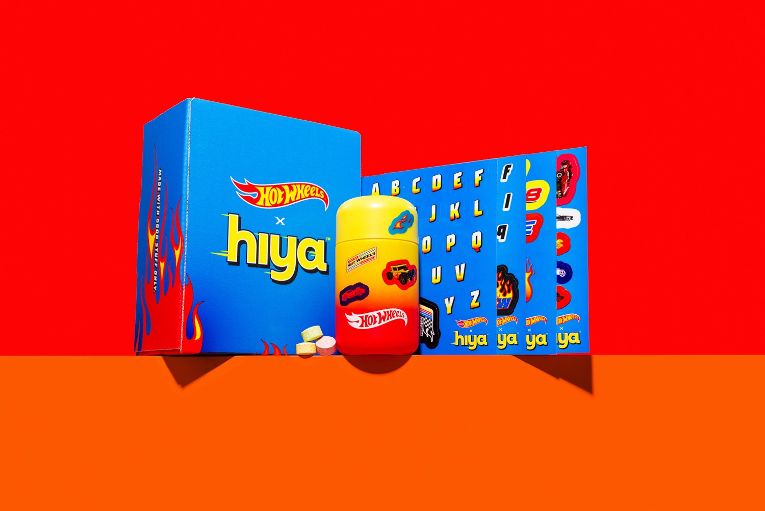Hot Wheels x Hiya collectibles packaging, featuring a bright yellow capsule with images of a red car and a black Jeep, alongside alphabet cards and Hot Wheels branding, on a vibrant red background with an orange surface.