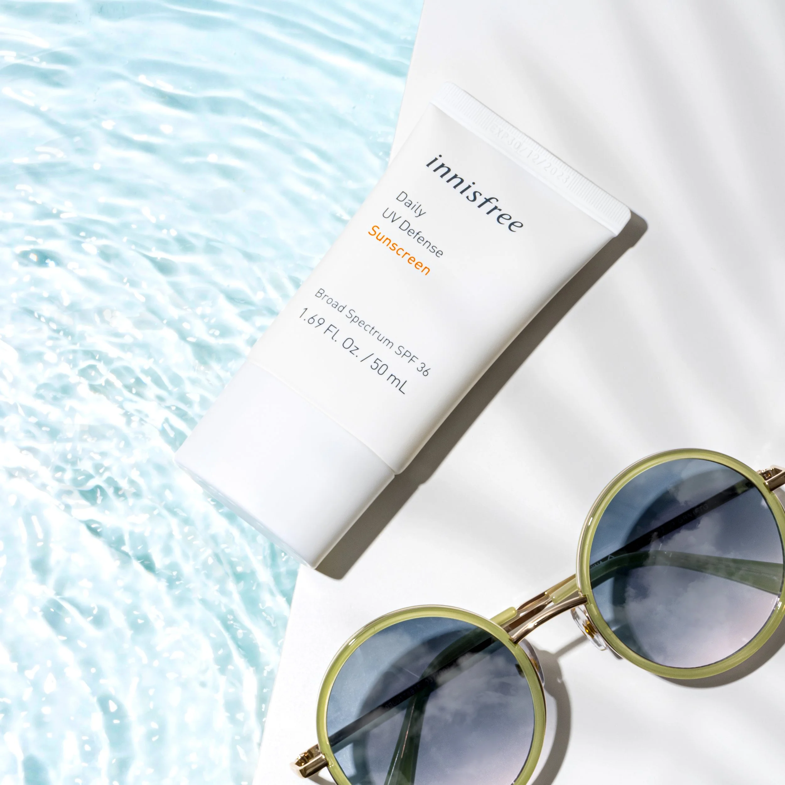 A tube of Innisfree Daily UV Defense Sunscreen with broad spectrum SPF 36, next to a pair of round, green sunglasses with tinted lenses, all on a white and blue water background.