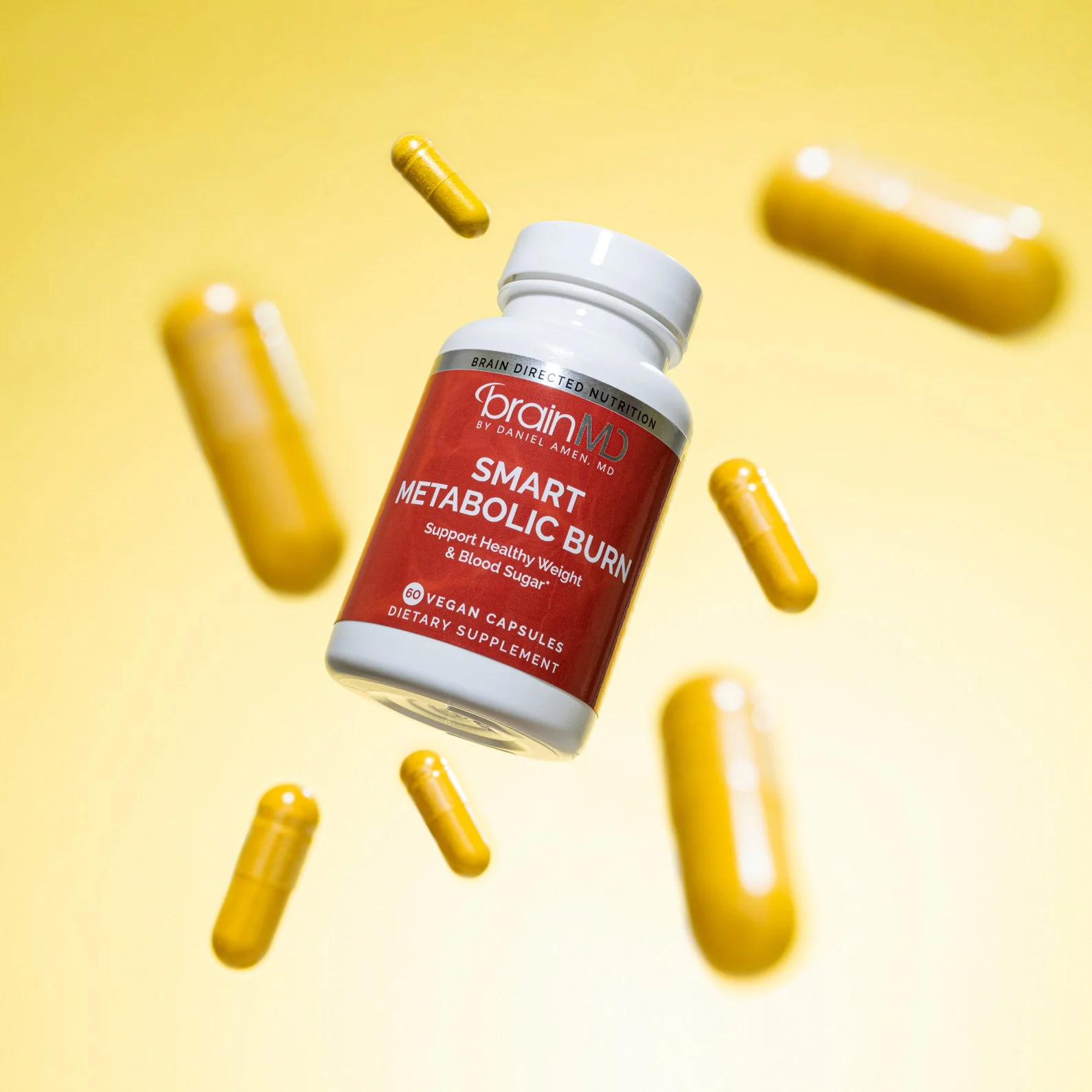 Bottle of Brain MD Smart Metabolic Burn dietary supplement with yellow capsules floating around it on a yellow background.