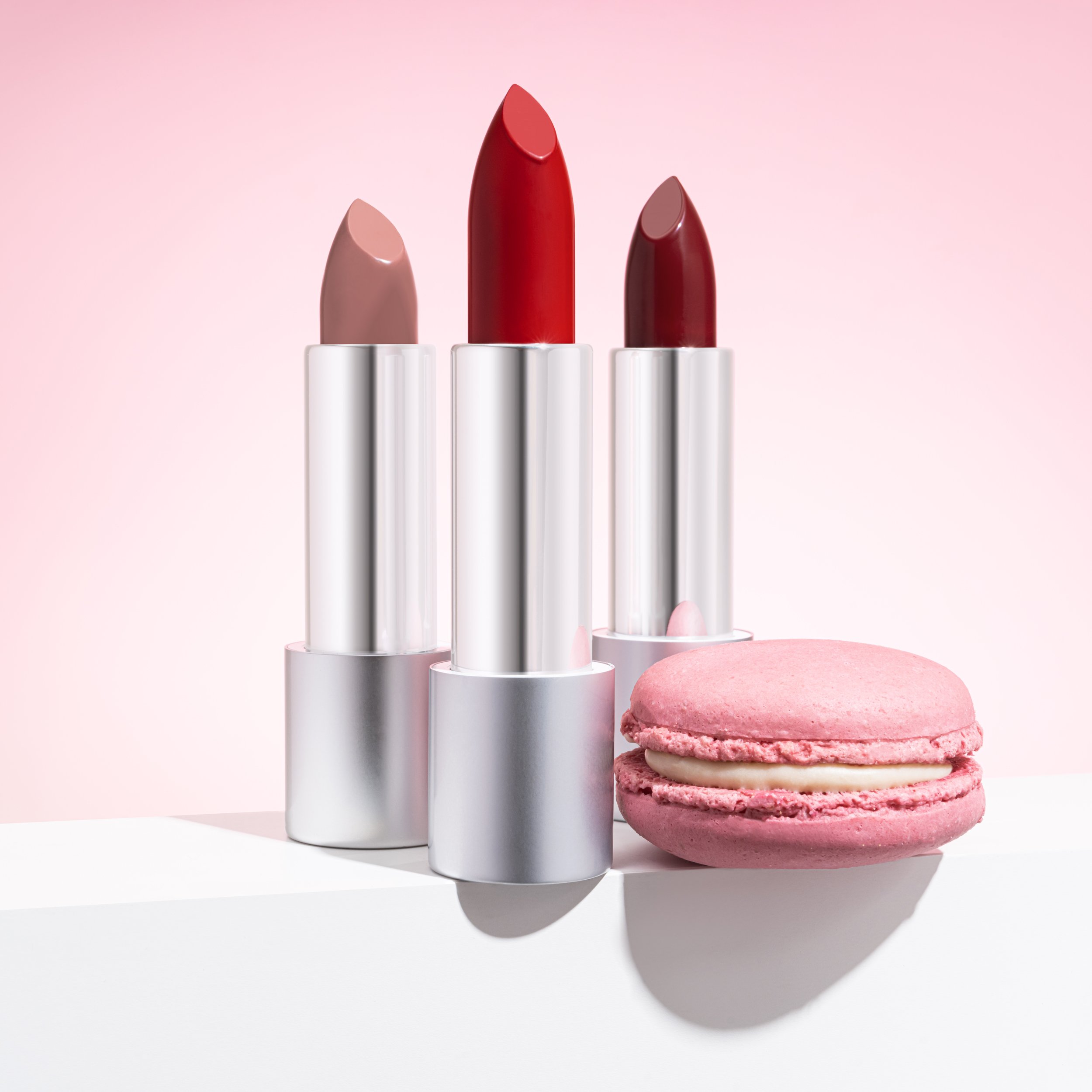 Three lipsticks in shades of nude and red and a pink macaron on a white surface against a light pink background.