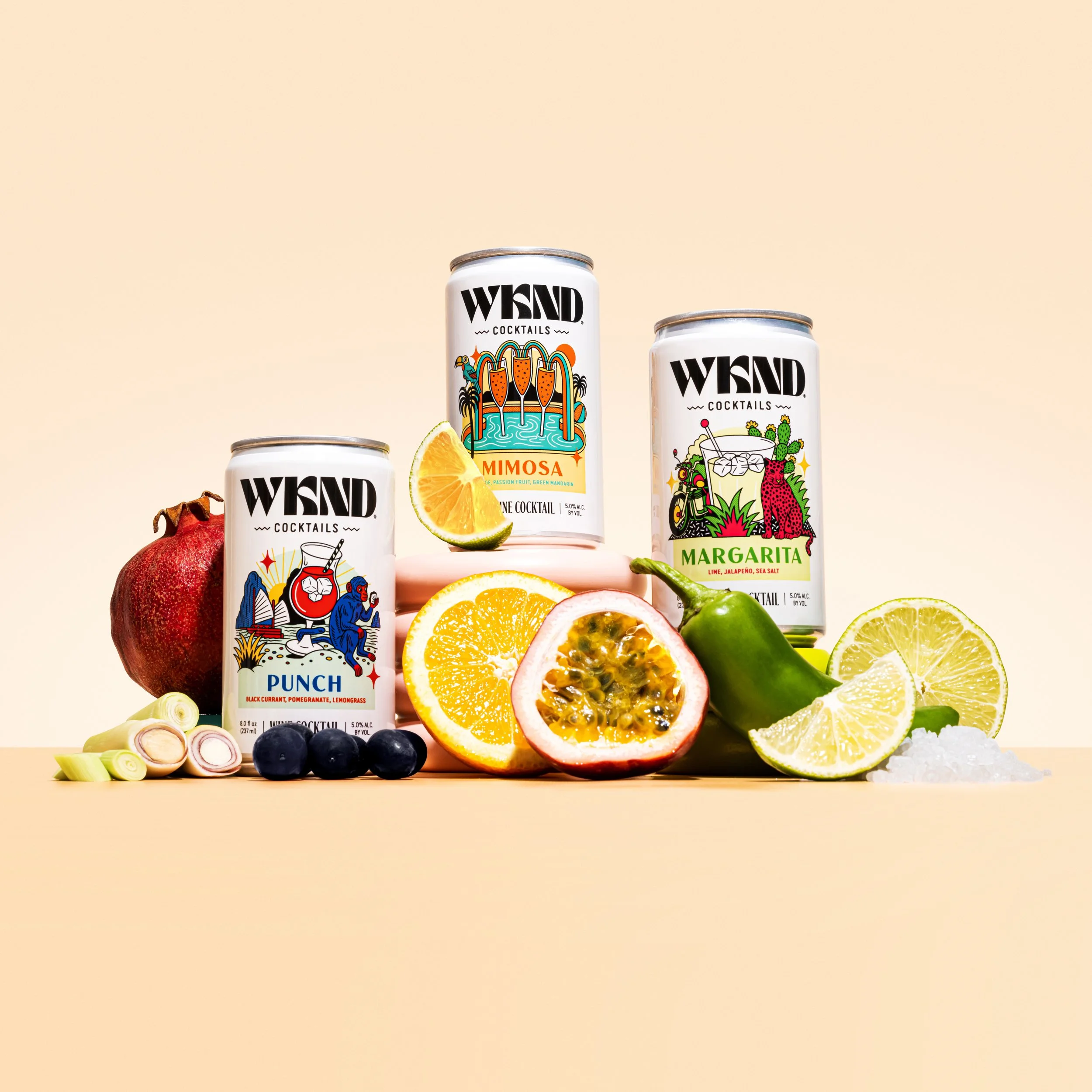 Three cans of WYLD cocktail beverages with fresh fruit slices, including red pomegranate, black blueberries, yellow lemon, green lime, green jalapeño, and a passion fruit, arranged on a beige background.